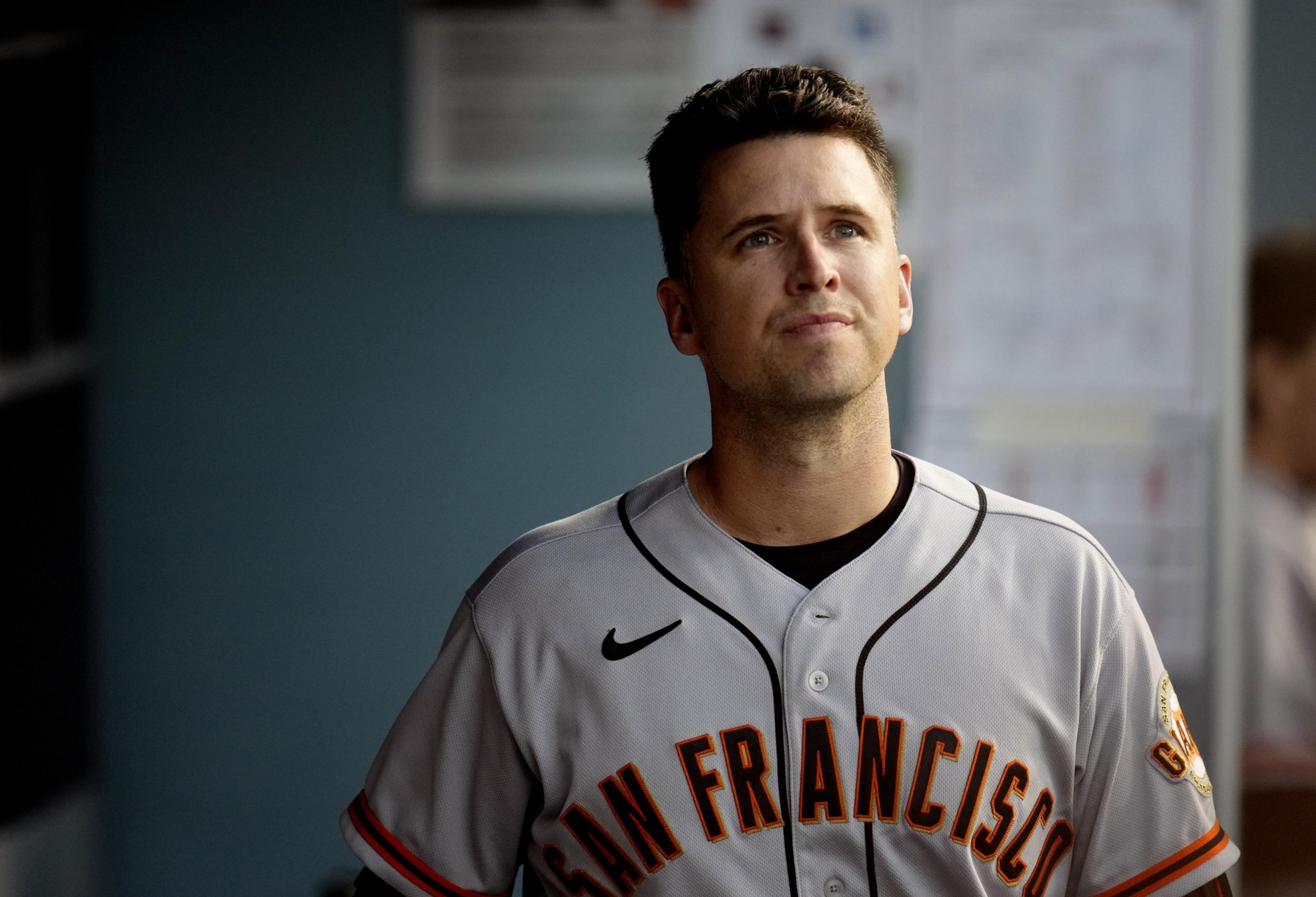 A baseball player in a San Francisco jersey looks upwards while standing in a dugout, with blurred background elements and natural lighting.