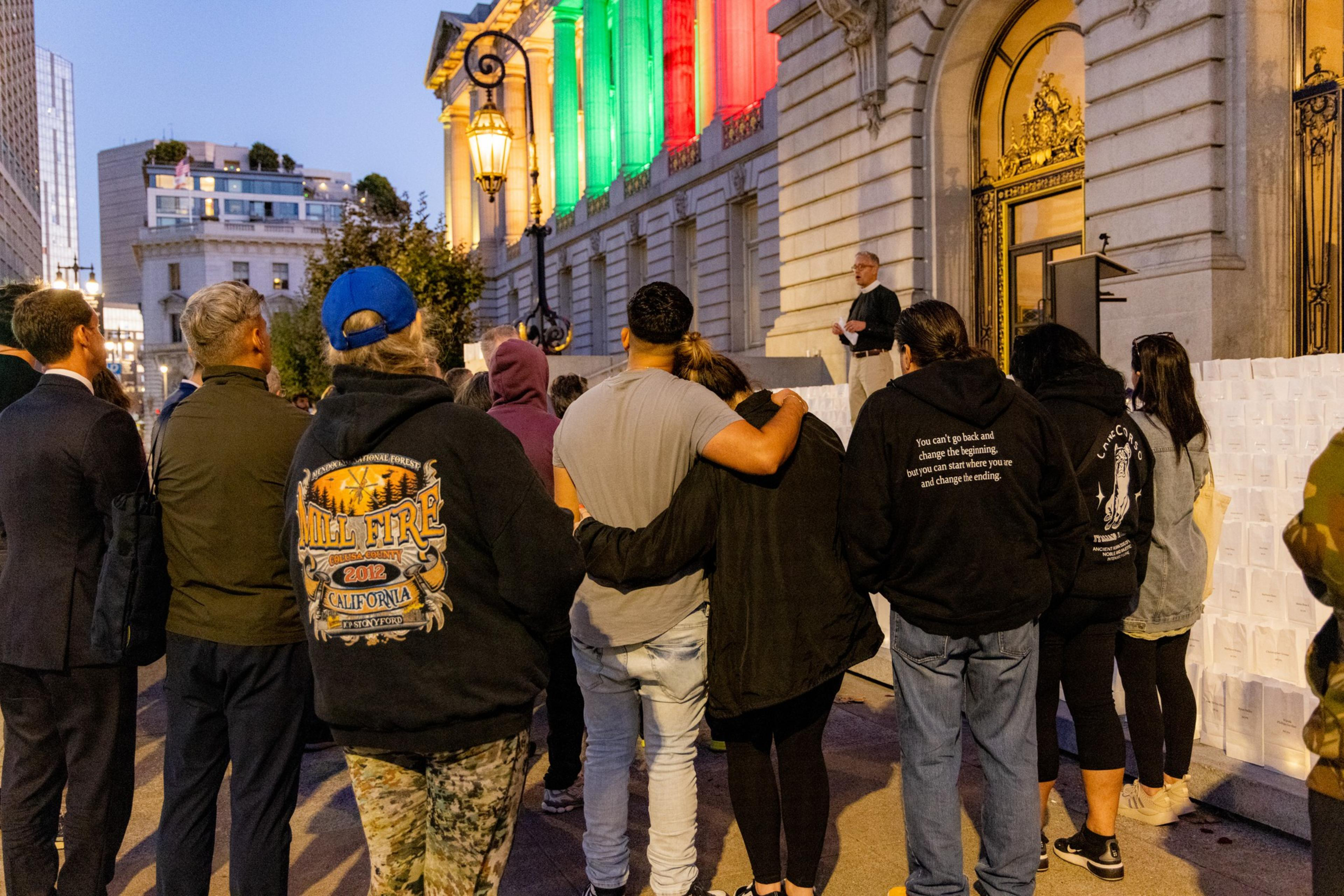 A group of people gather outside a building lit with green and red lights, watching a speaker. Some individuals are hugging, creating a sense of community.