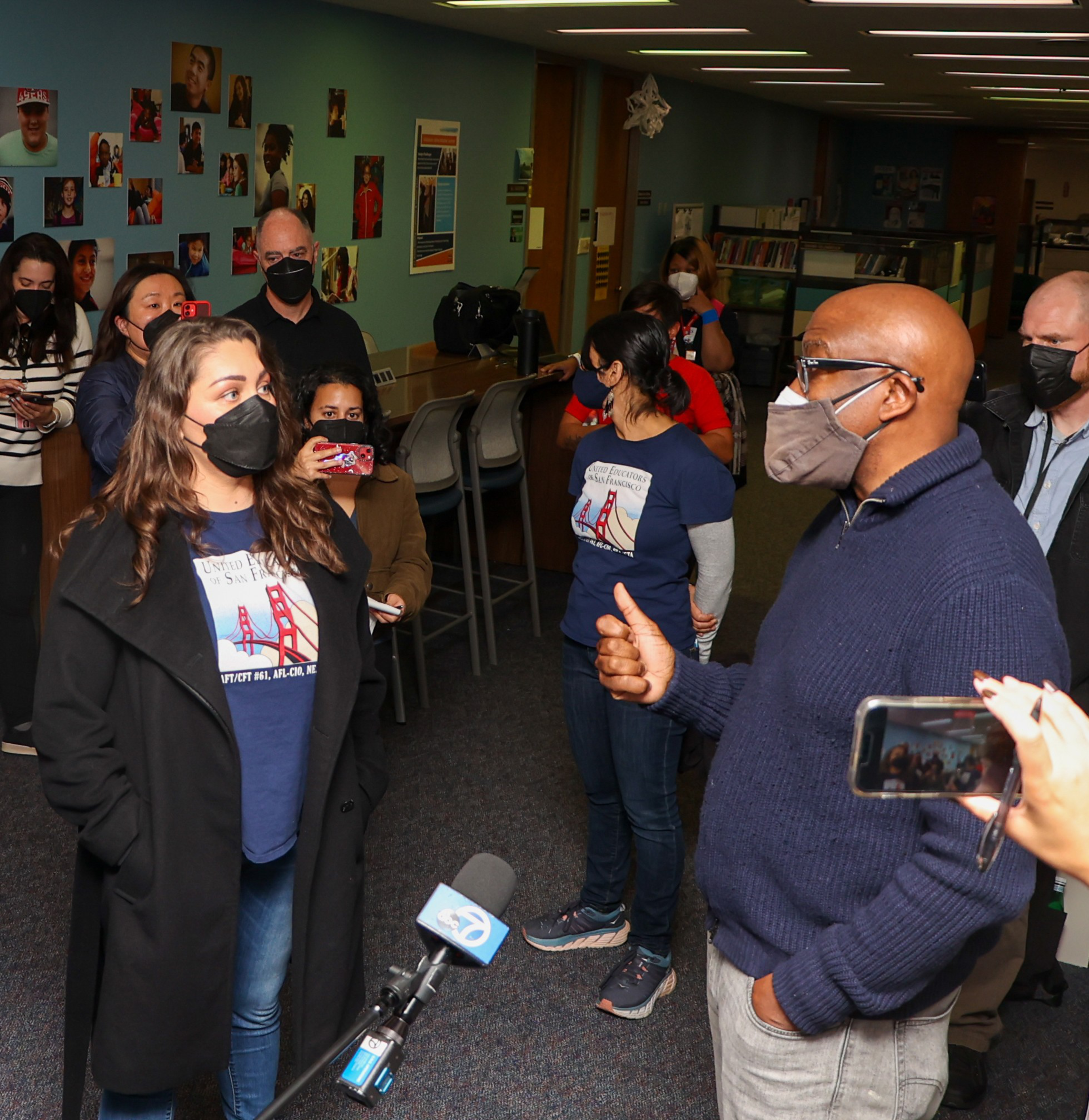 A group of masked people stand indoors, two in matching navy shirts with a Bridge graphic talk while others listen and record with phones and cameras.