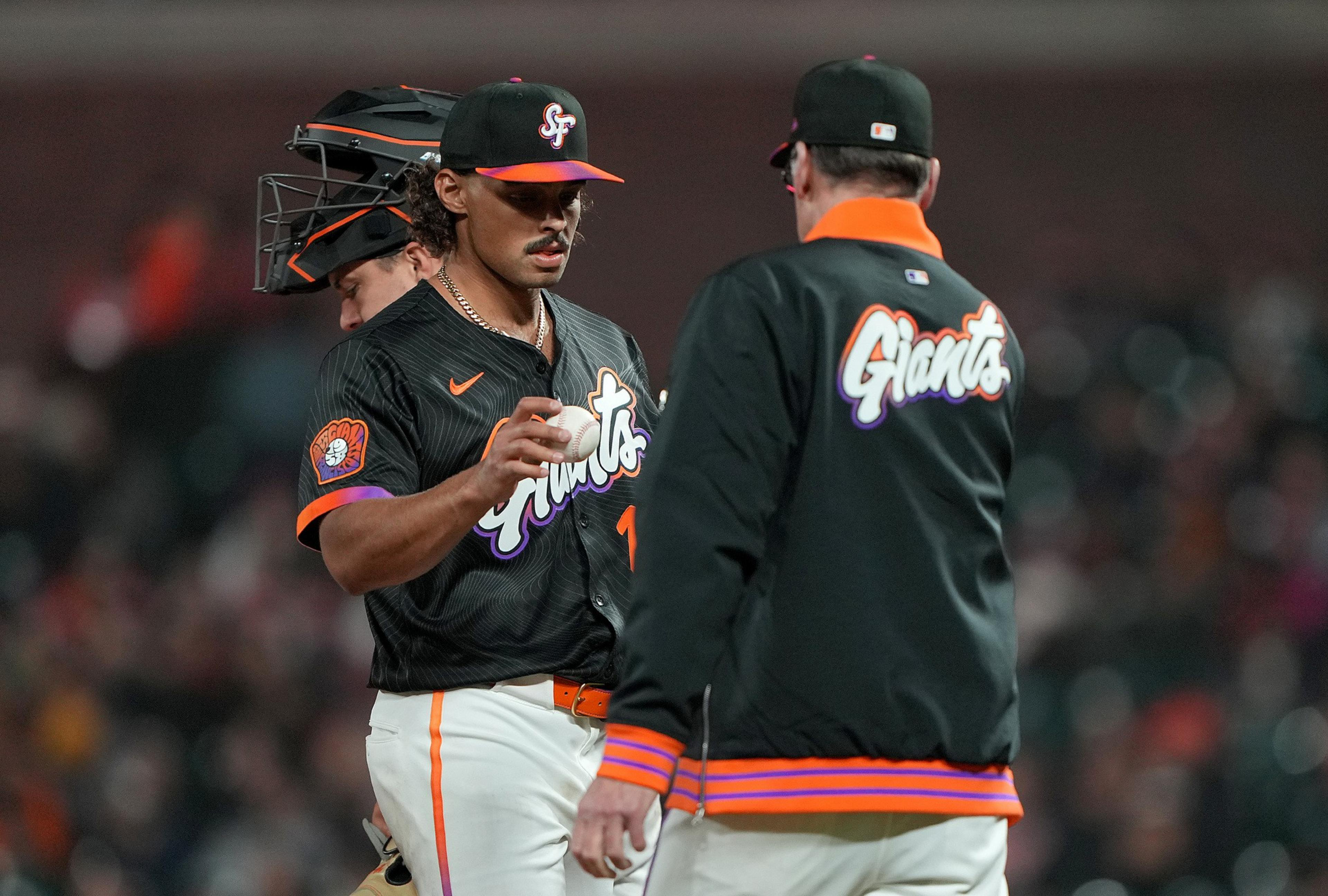 A baseball player in a black Giants uniform with colorful details holds a baseball, talking to a coach. A catcher with a helmet stands nearby.