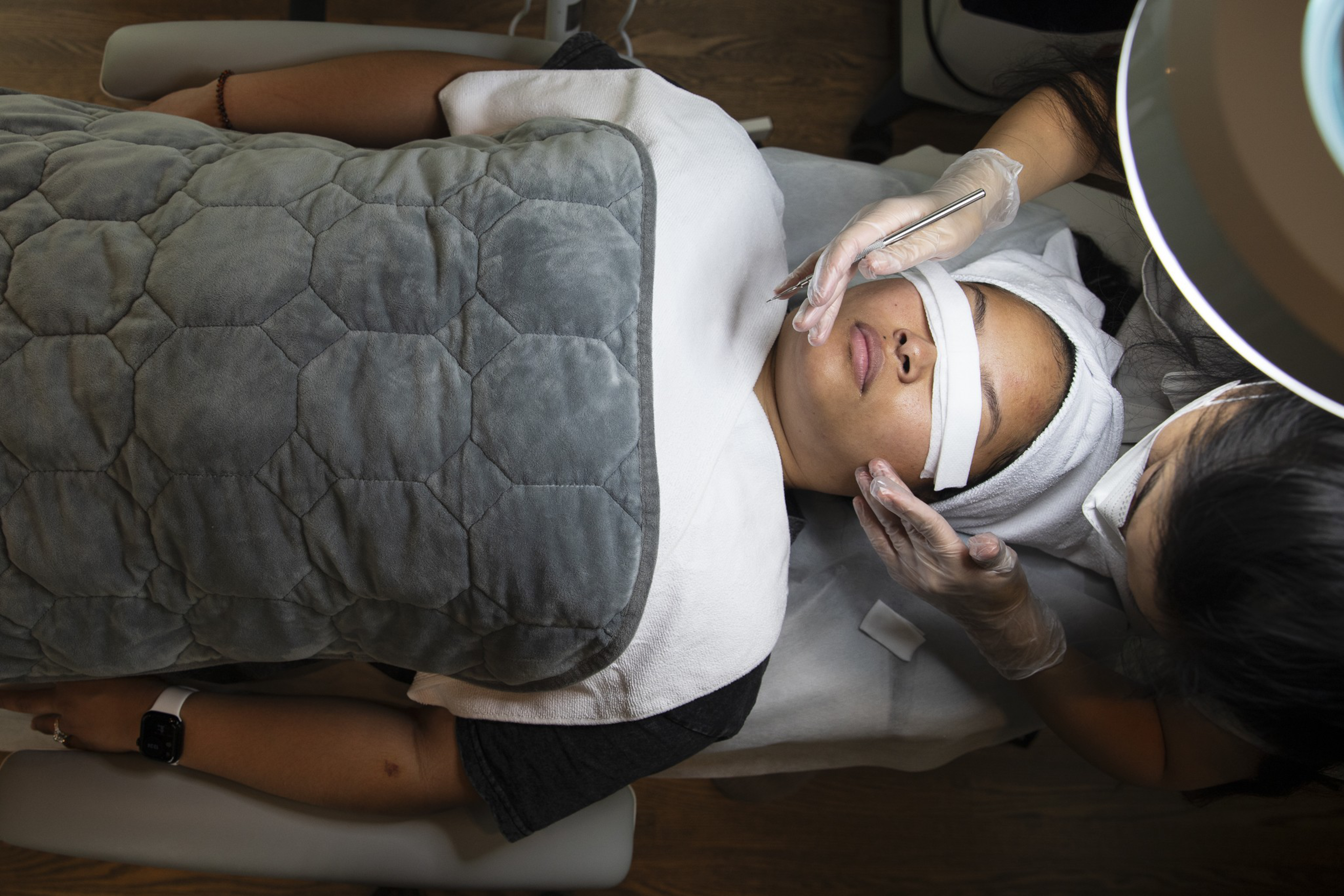 A person lies on a treatment bed with eyes covered, receiving a facial procedure from a gloved professional using a metal tool under a bright ring light.