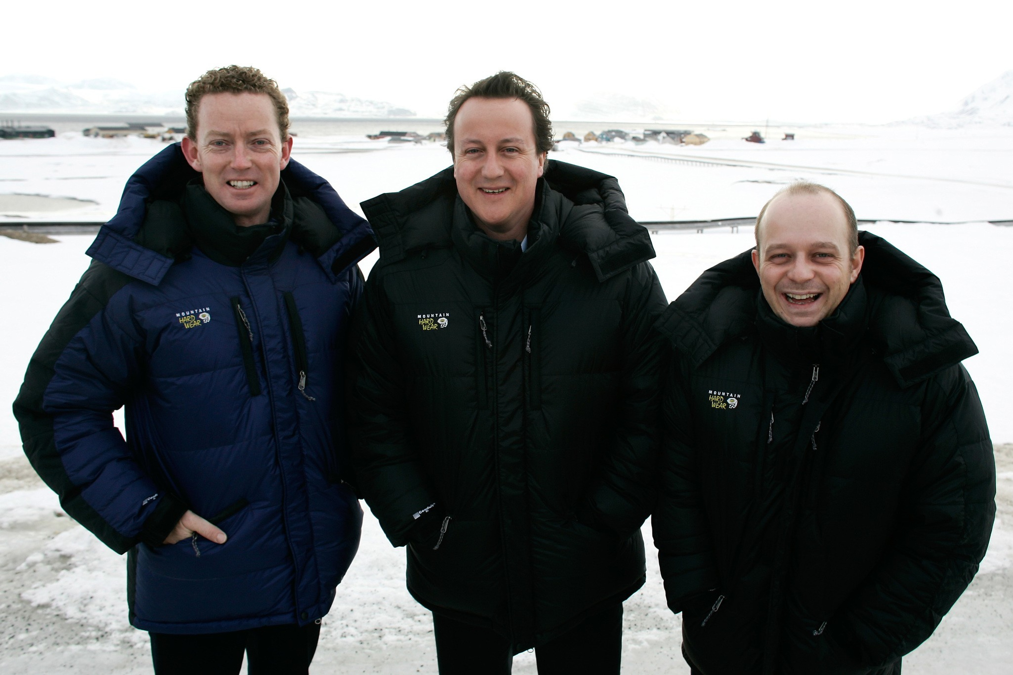 Three men wearing thick winter jackets stand outdoors on snow-covered ground, smiling with a snowy landscape and distant mountains behind them.