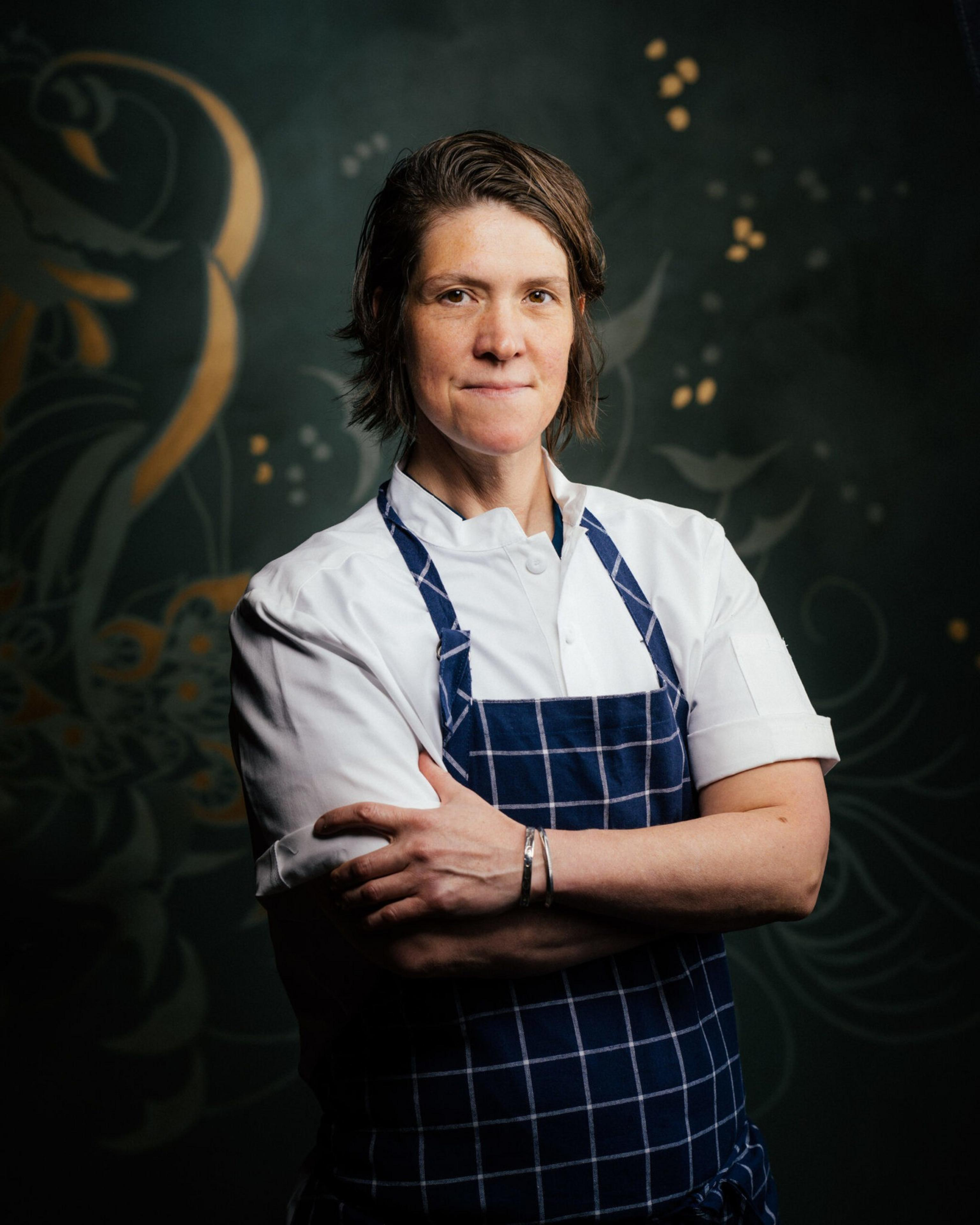 A chef in a blue apron stands with her arms crossed.