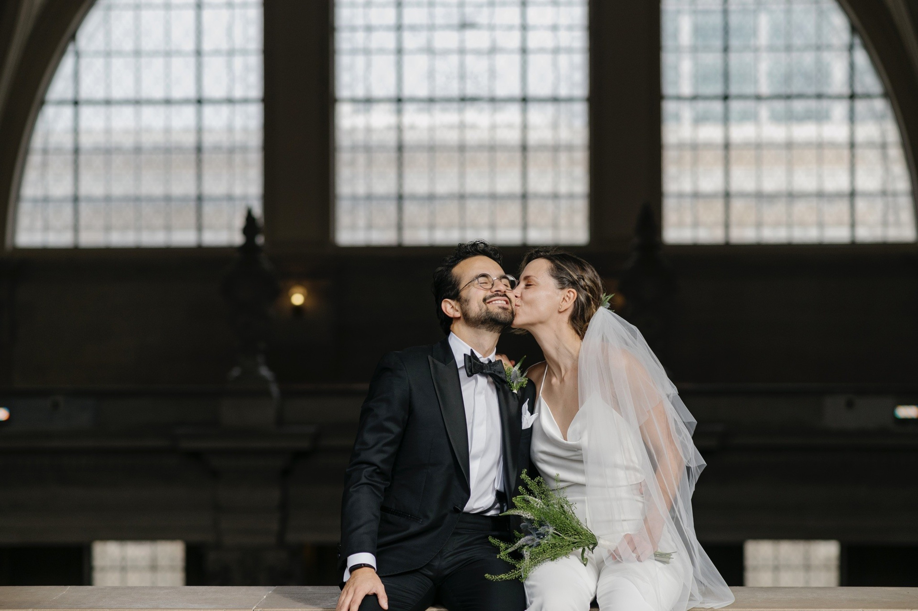 A wedding couple, seated, kisses in front of a large window. The person on the left wears a suit and bow tie, the one on the right wears a white pantsuit and veil.