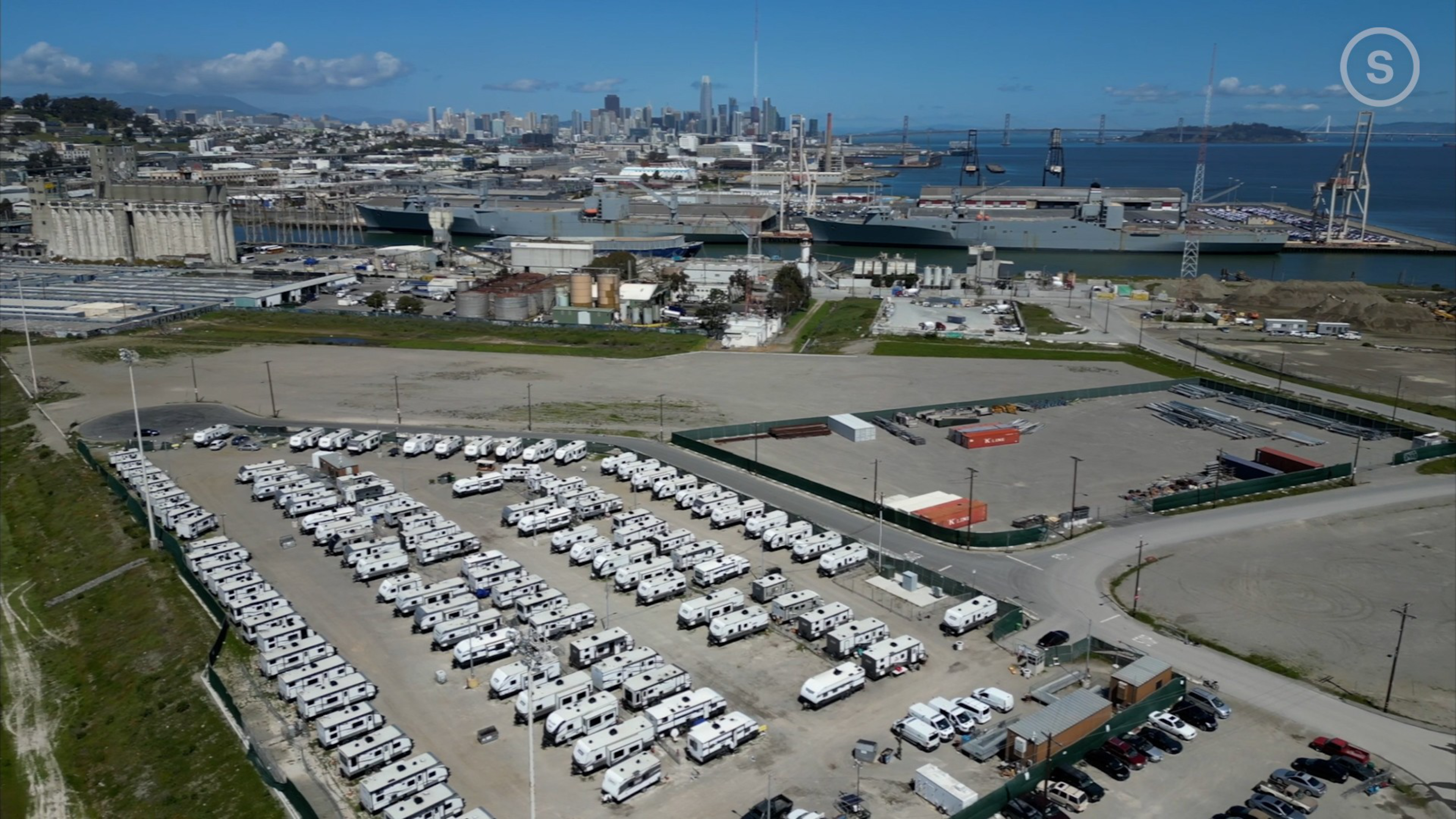A large parking lot filled with rows of white recreational vehicles, an aircraft carrier in the harbor, and a city skyline in the background under a clear blue sky.
