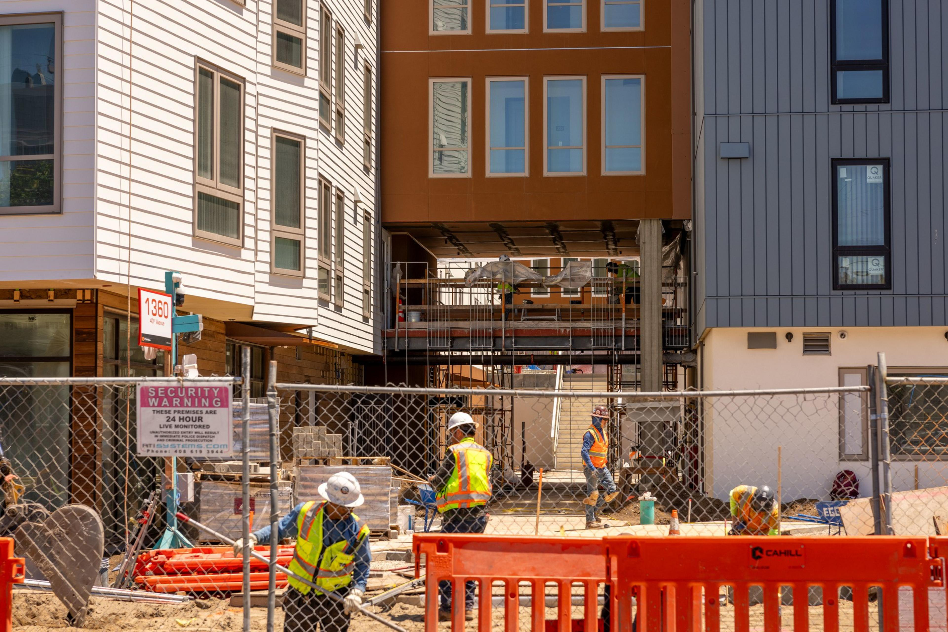 Construction workers in safety gear work at a site between modern buildings. Fences and signs are visible, with various construction materials around.