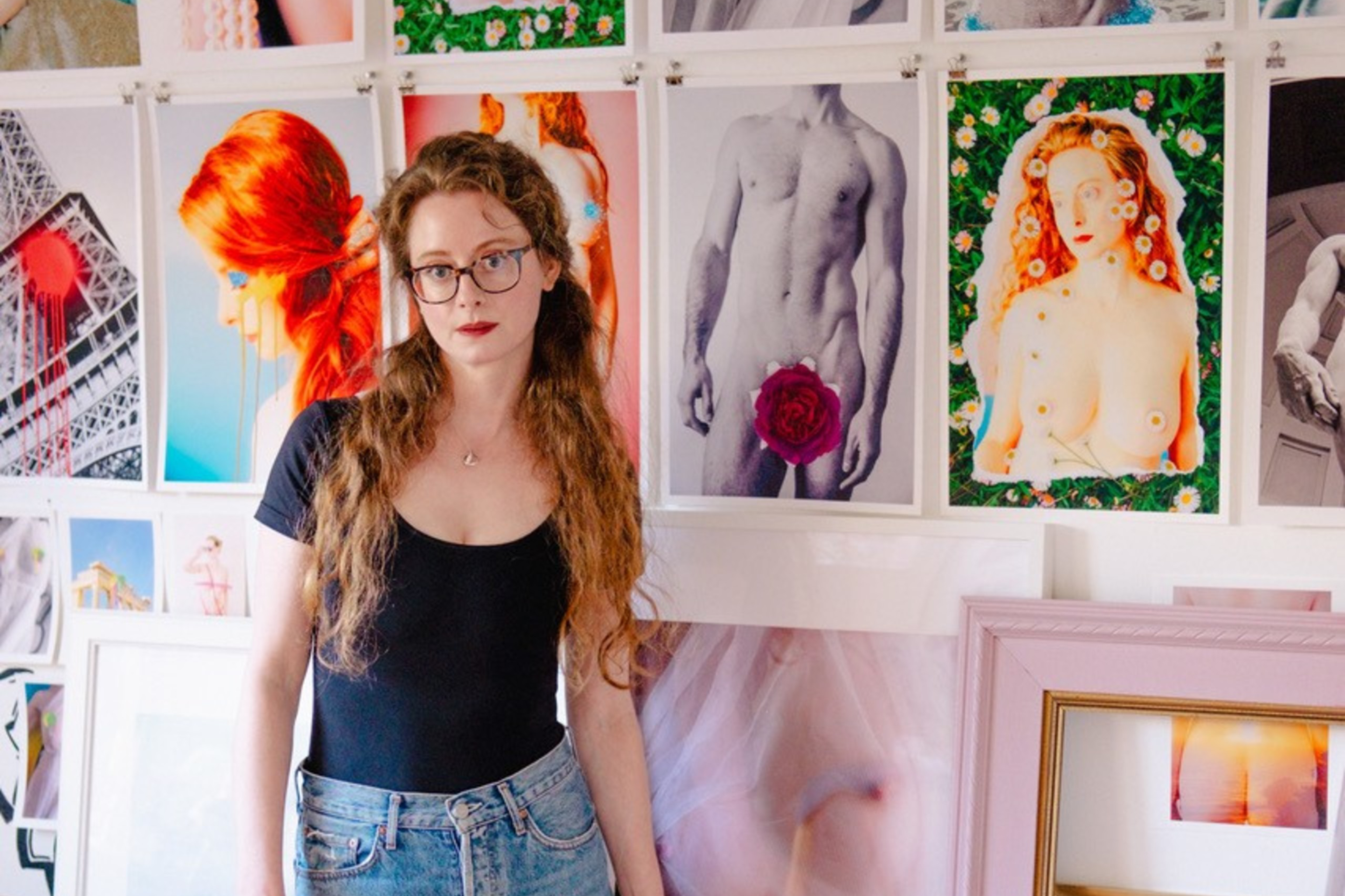 A woman with glasses, wearing a black top and ripped jeans, stands barefoot in front of a wall filled with vibrant photographic prints, mostly of artistic nudes.