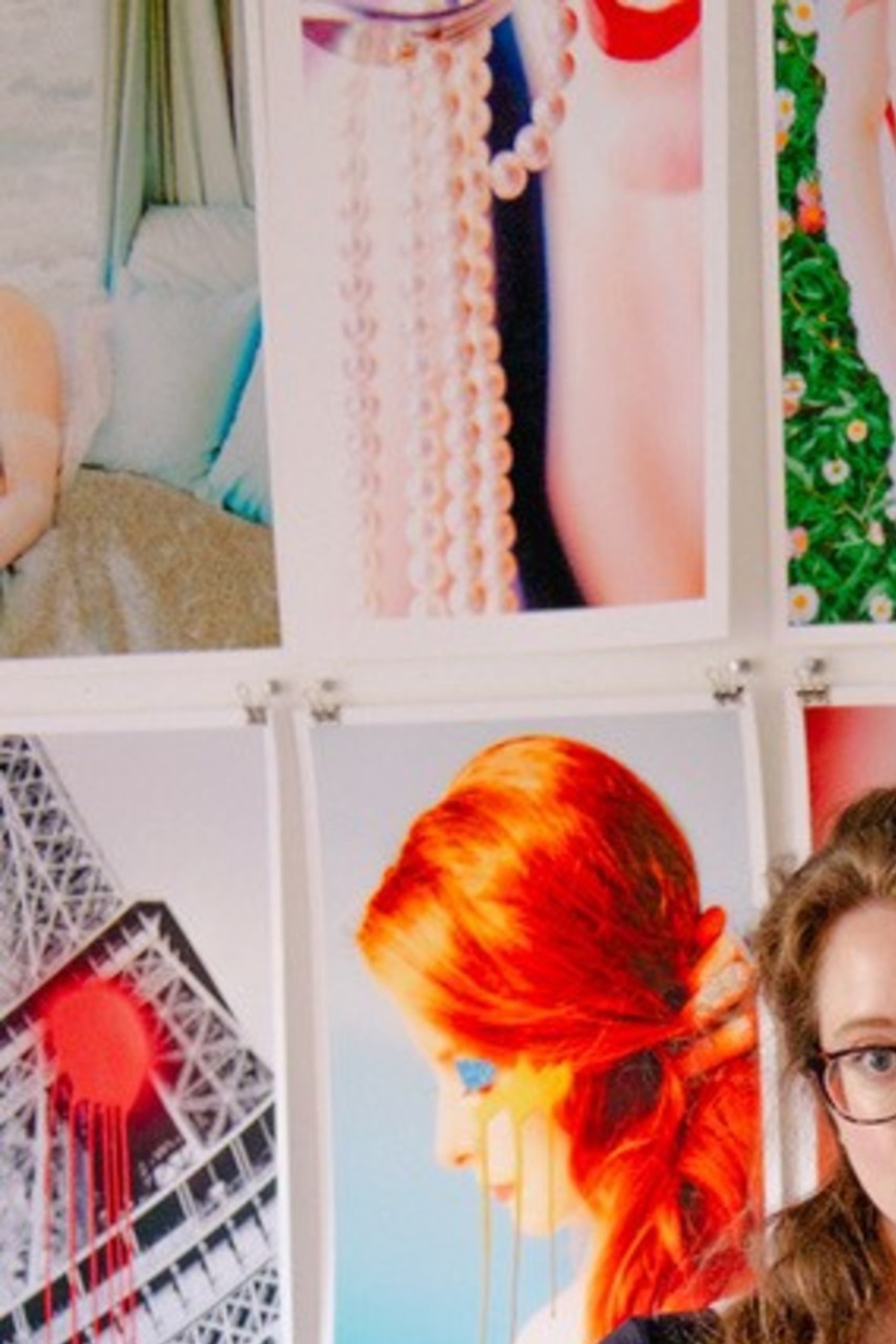 A woman with glasses, wearing a black top and ripped jeans, stands barefoot in front of a wall filled with vibrant photographic prints, mostly of artistic nudes.