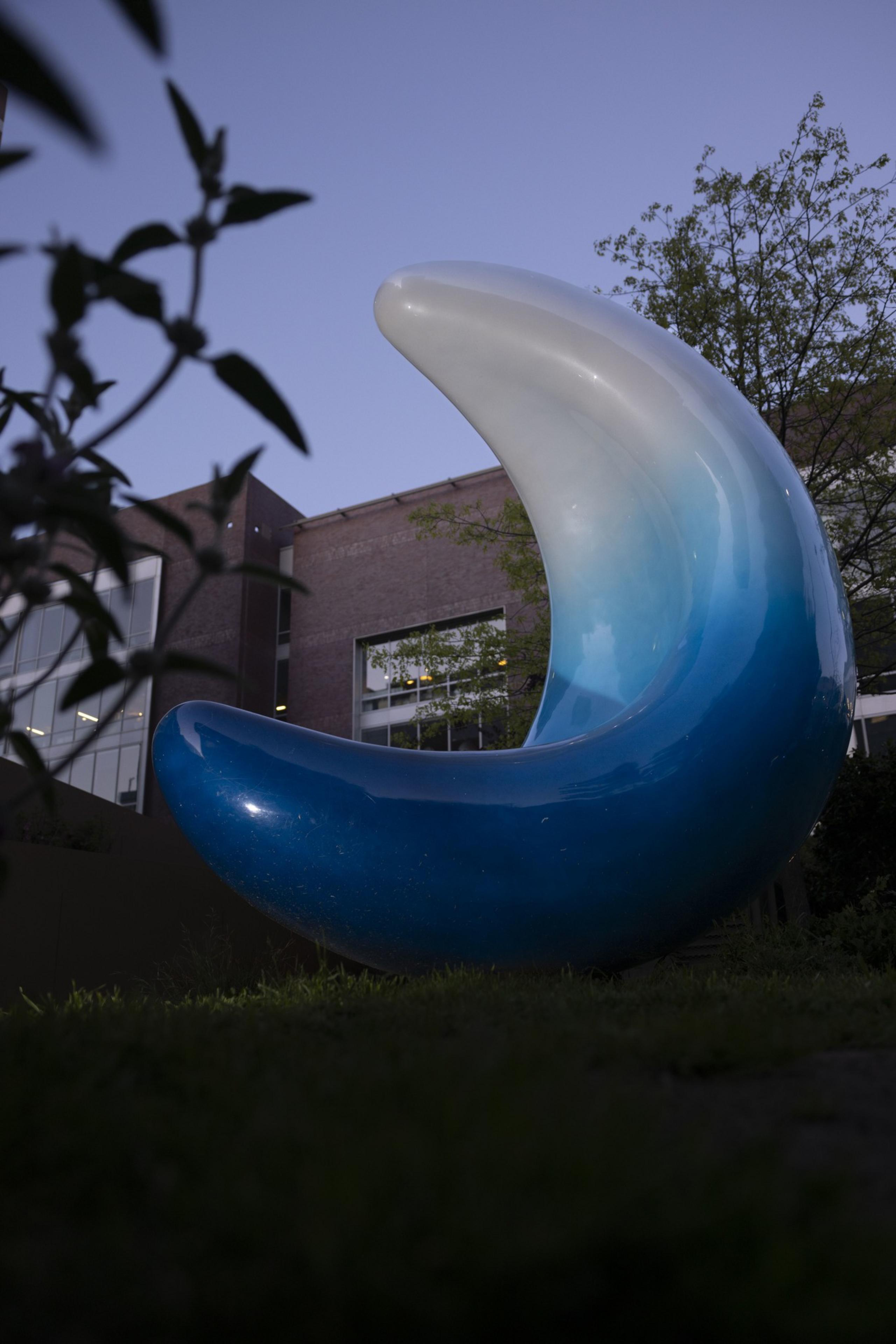 A large blue and white crescent sculpture stands outdoors, surrounded by grass and plants, with a building and trees visible in the background.