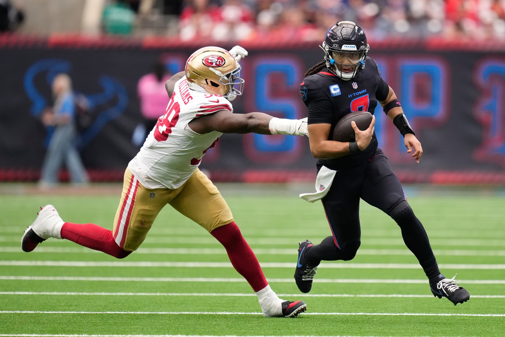 A football player in a black uniform runs with the ball while a player in a white and gold uniform lunges to tackle him.