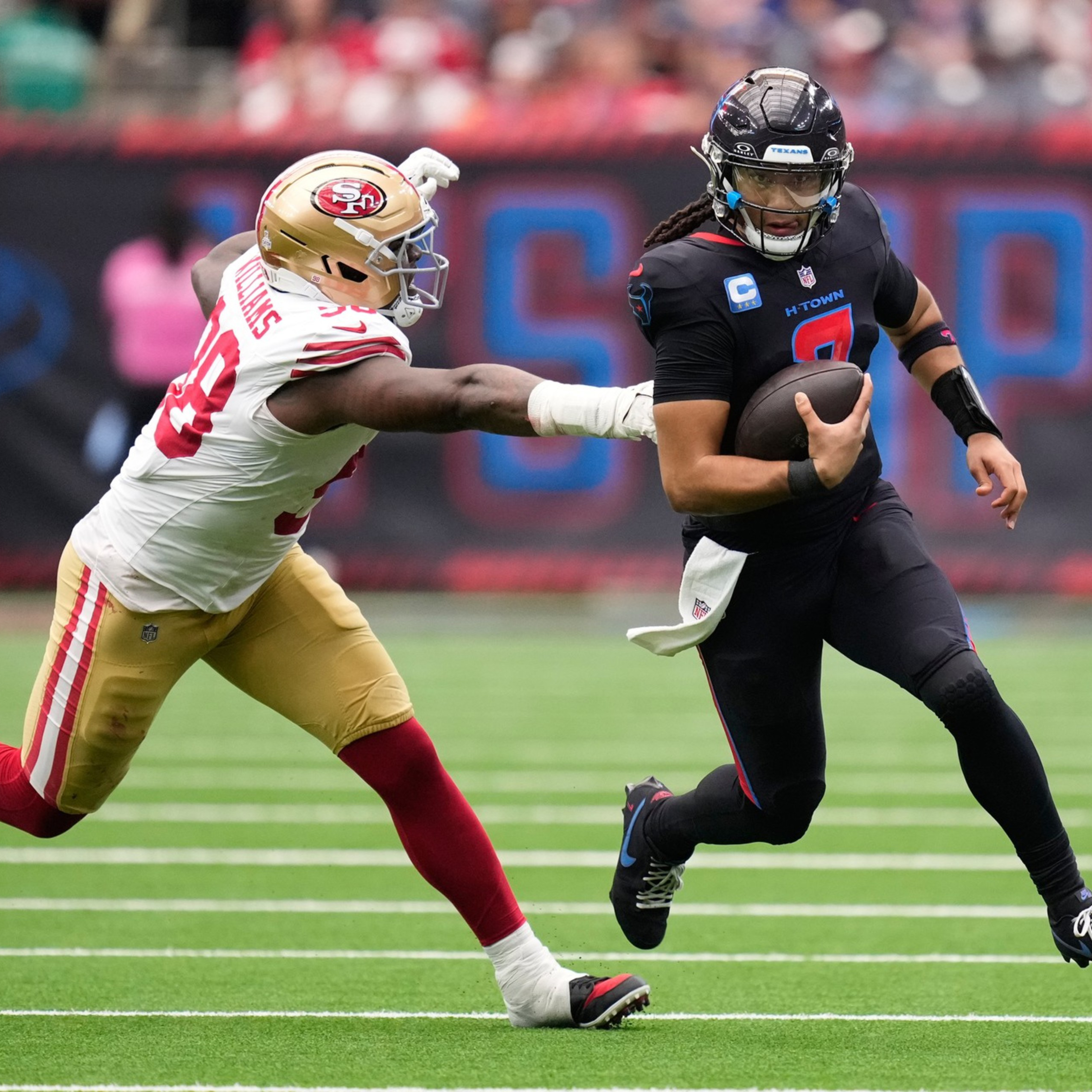 A football player in a black uniform runs with the ball while a player in a white and gold uniform lunges to tackle him.
