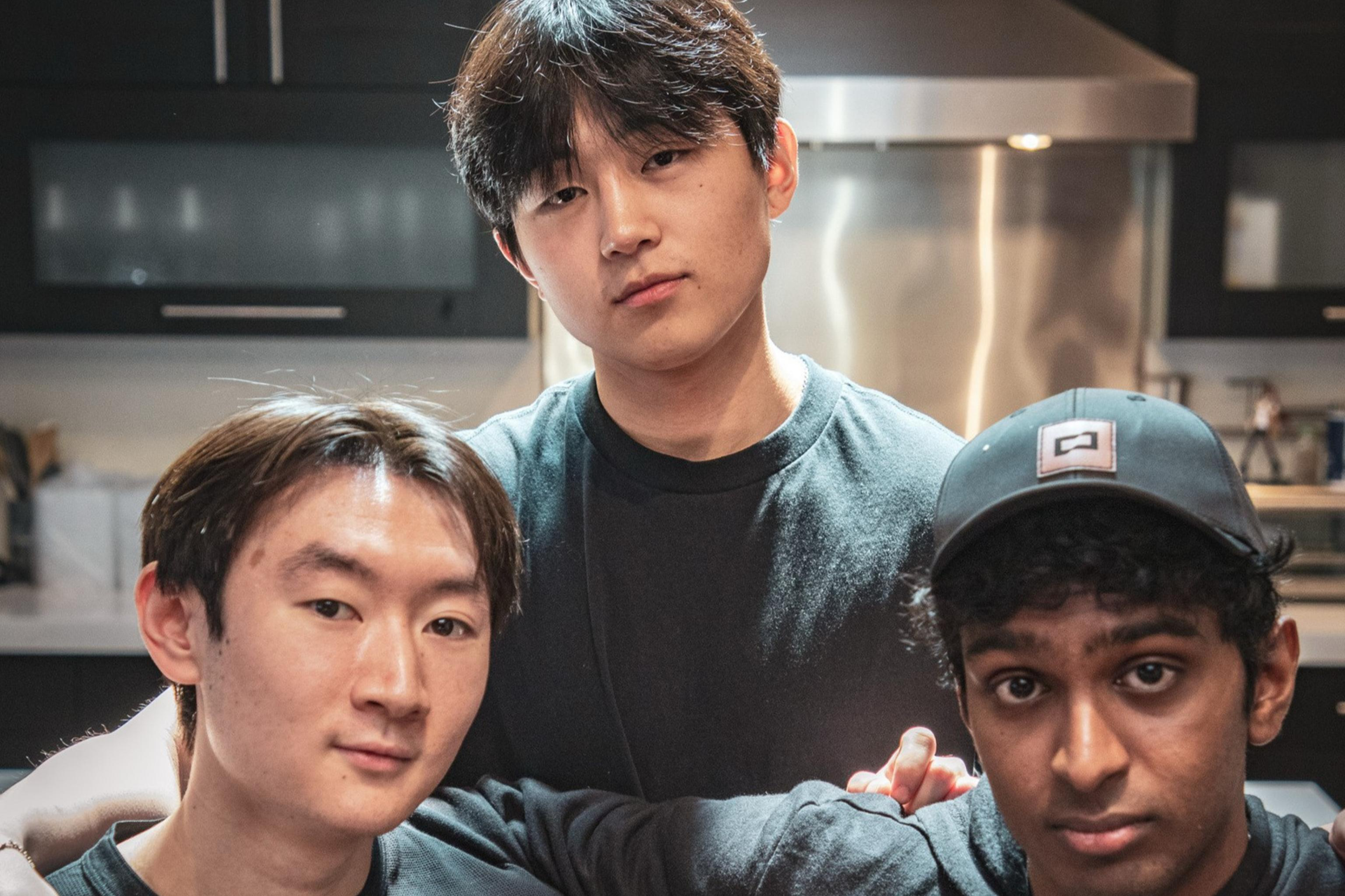 Three young men, wearing dark shirts, pose together in a kitchen. One stands while the other two sit, with arms around each other, under soft lighting.