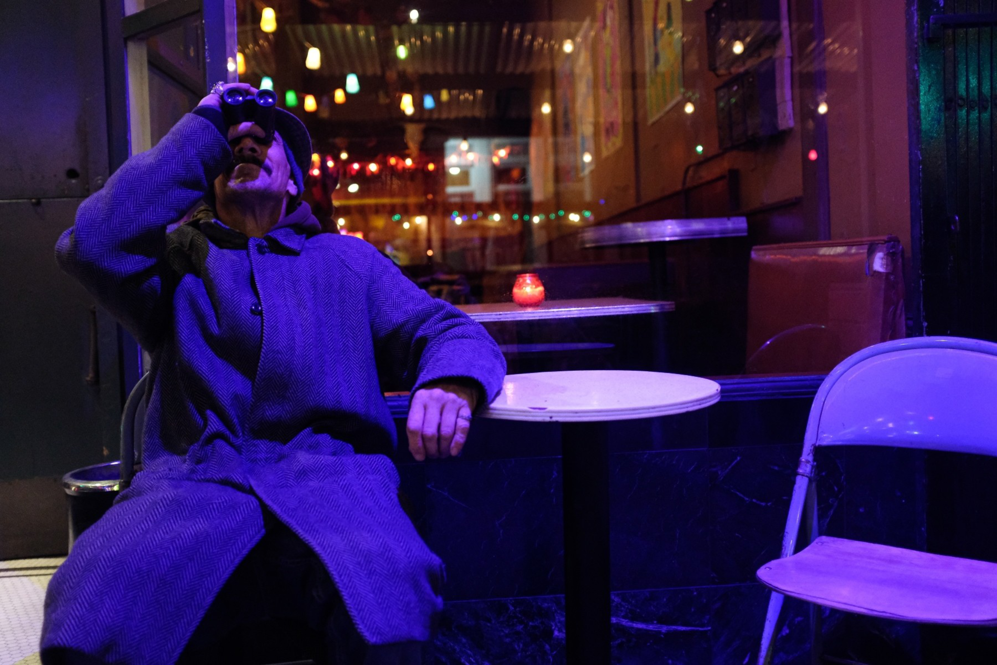 A person in a long coat and hat sits outside a dimly lit café at night, looking through binoculars with colorful lights reflected in the window.