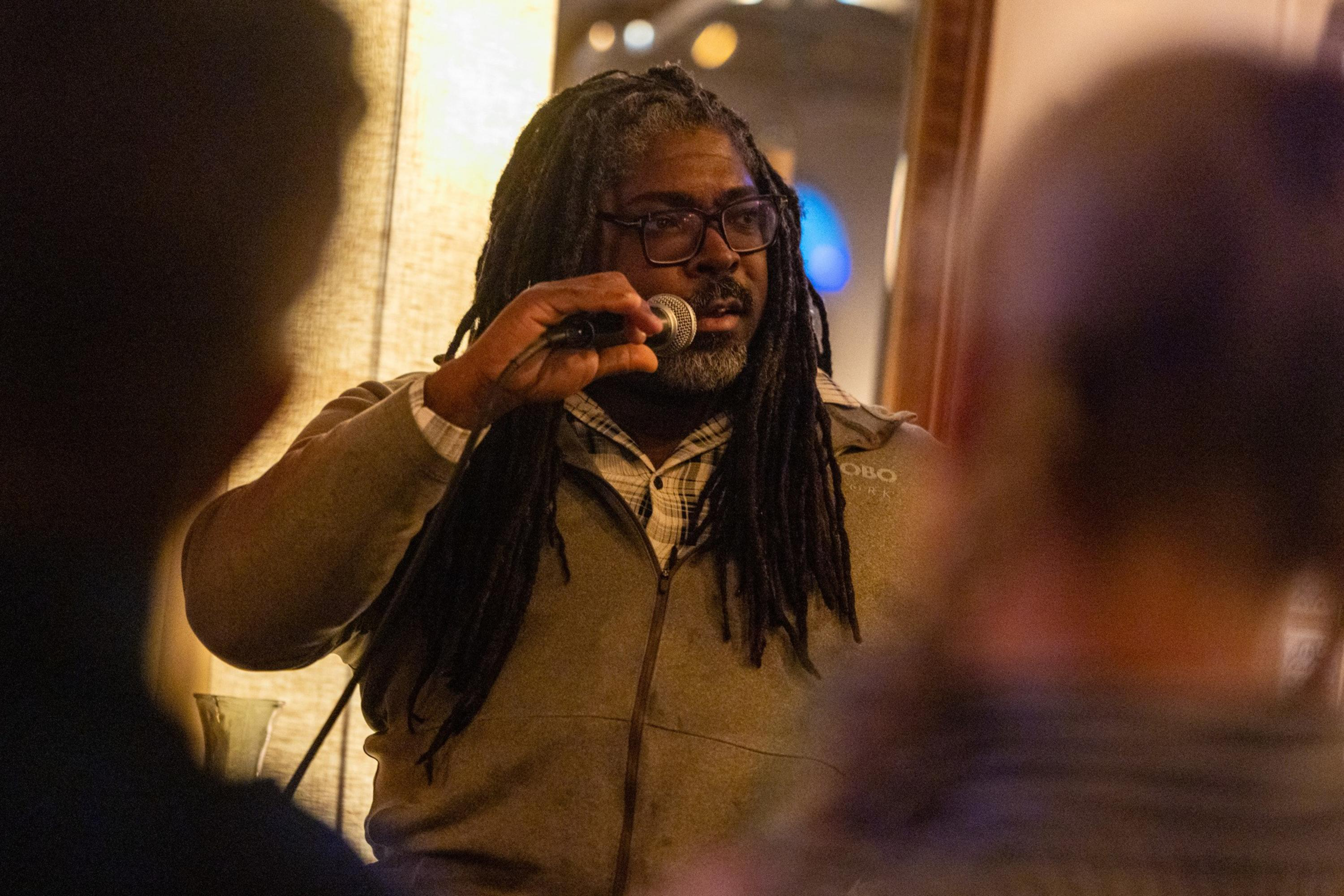 A man with long dreadlocks and glasses speaks into a microphone, looking to his left, while two blurred figures face him in the foreground.