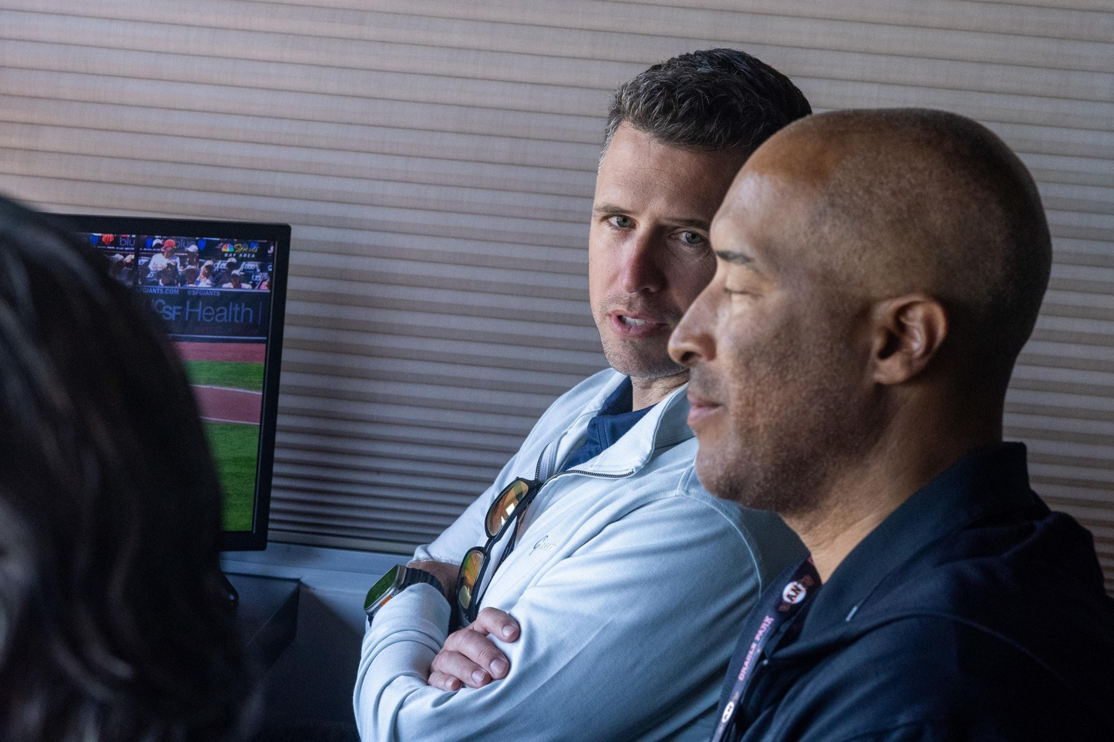 Two men are having a conversation in front of a monitor displaying a baseball game. One has short hair, wearing a light jacket with sunglasses on the collar.