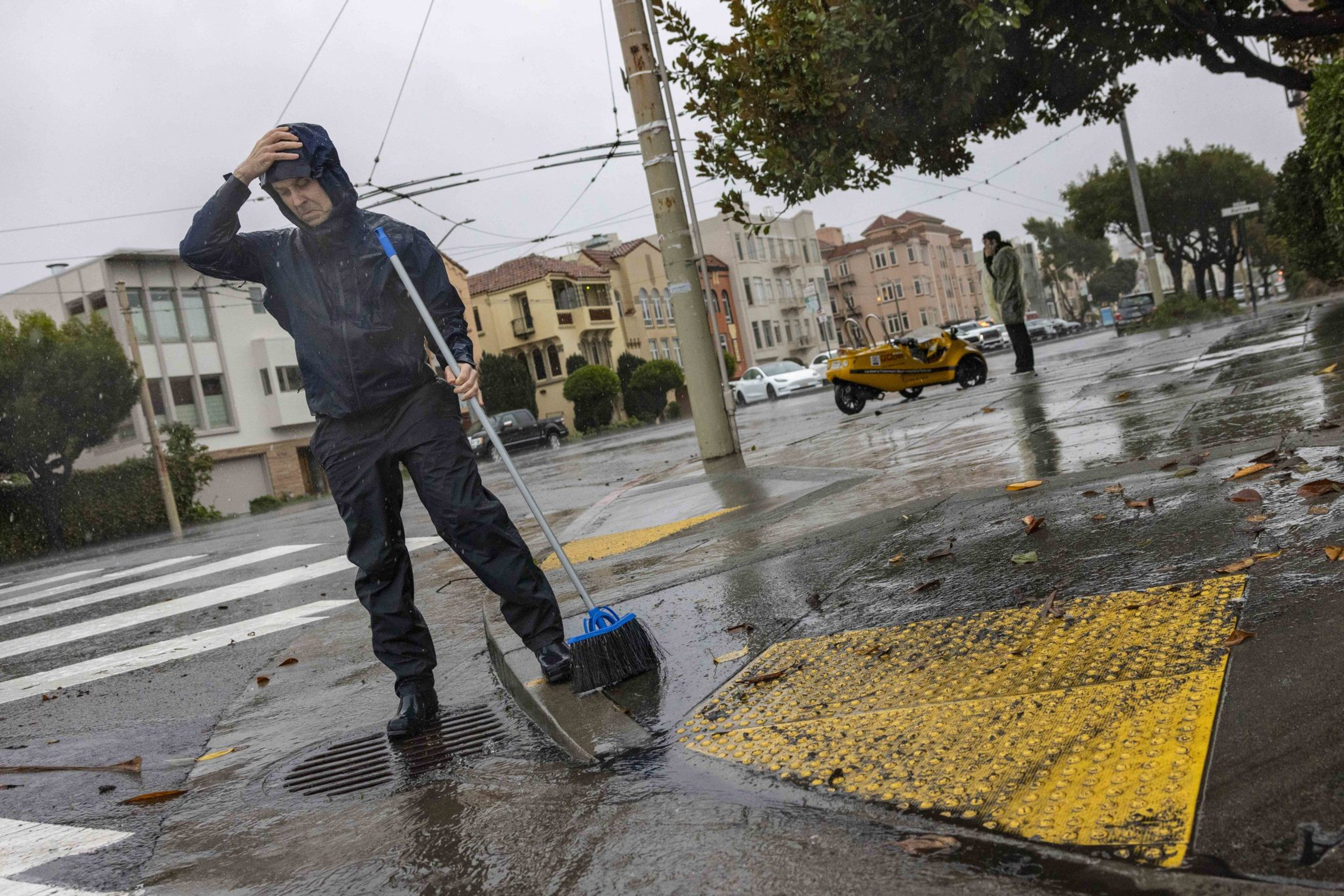 A person in rain gear sweeps water into a drain on a wet sidewalk, while holding a hood against the rain in a cityscape with buildings and a tree.