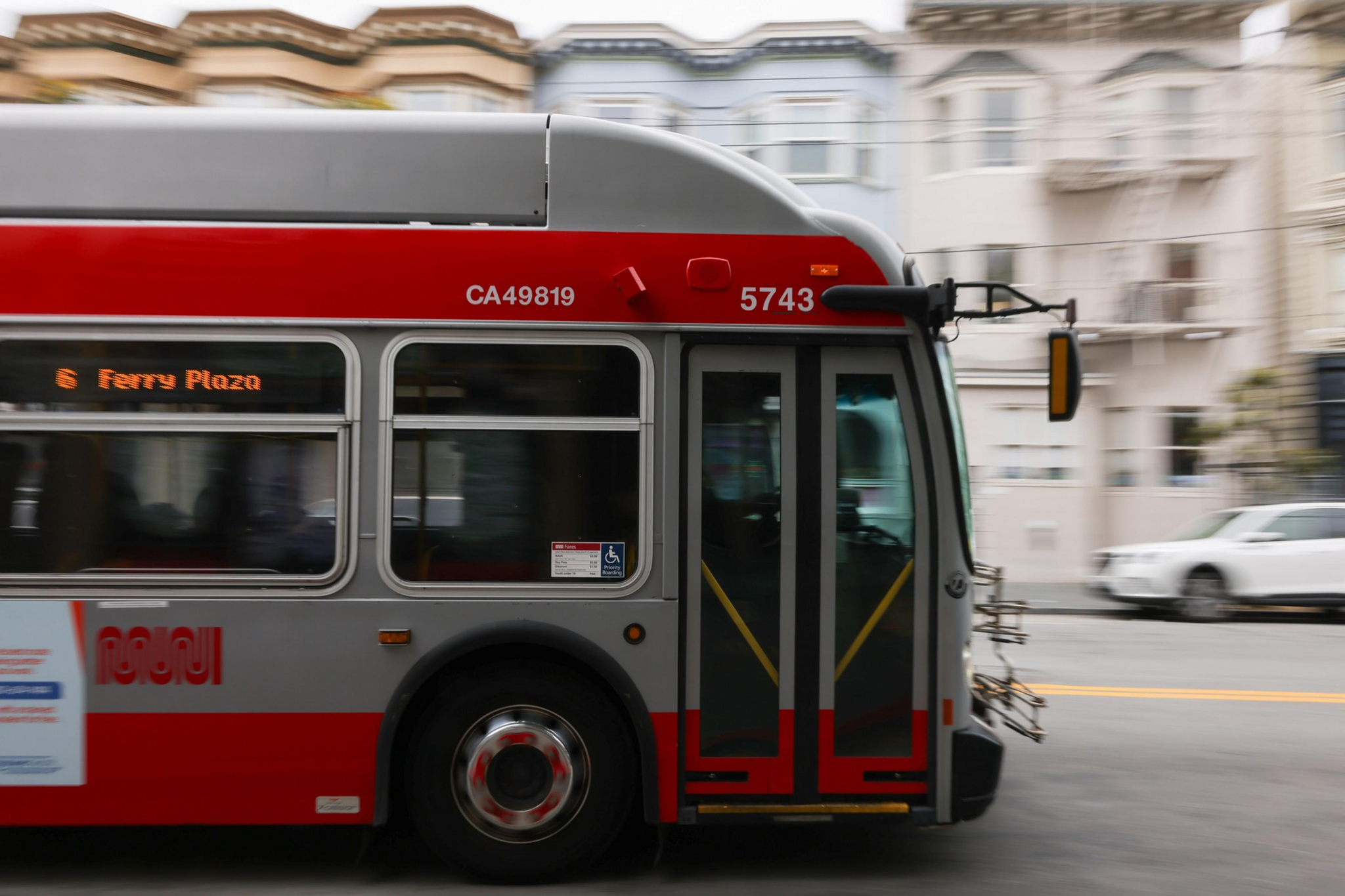 A red and gray city bus marked 5743 is in motion on a street with blurred buildings and cars in the background, displaying &quot;Ferry Plaza&quot; as its destination.