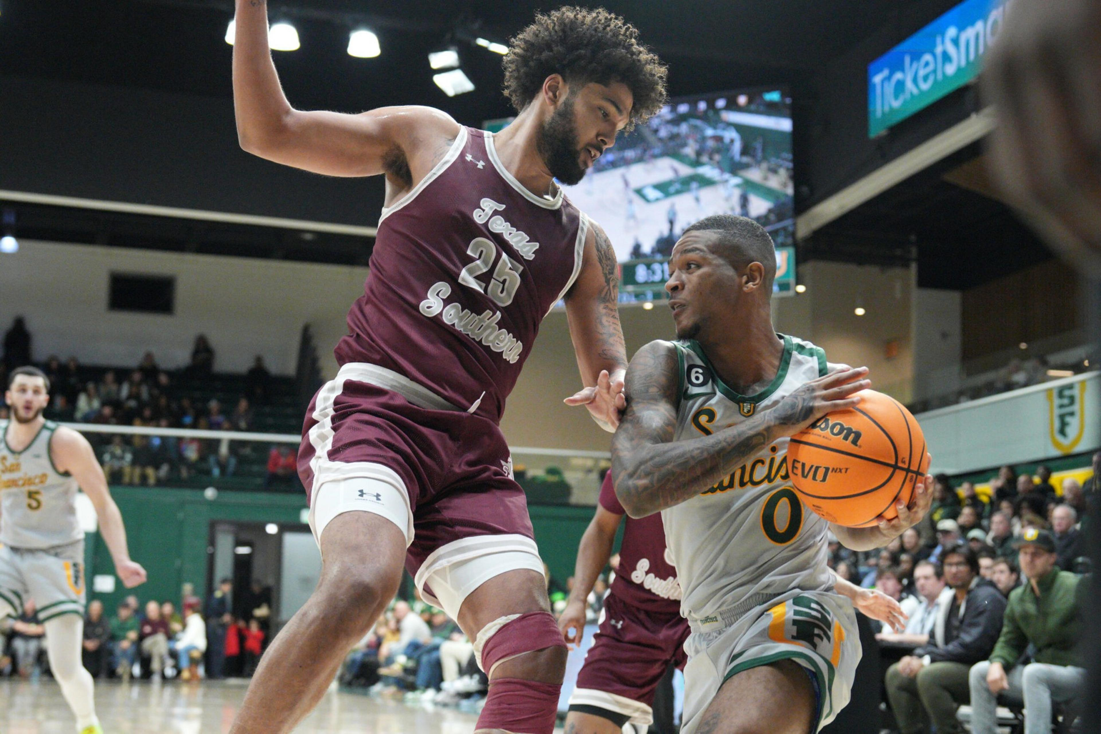 A basketball player in a gray and green uniform holds the ball while being closely guarded by a player in a maroon and white uniform during a game.