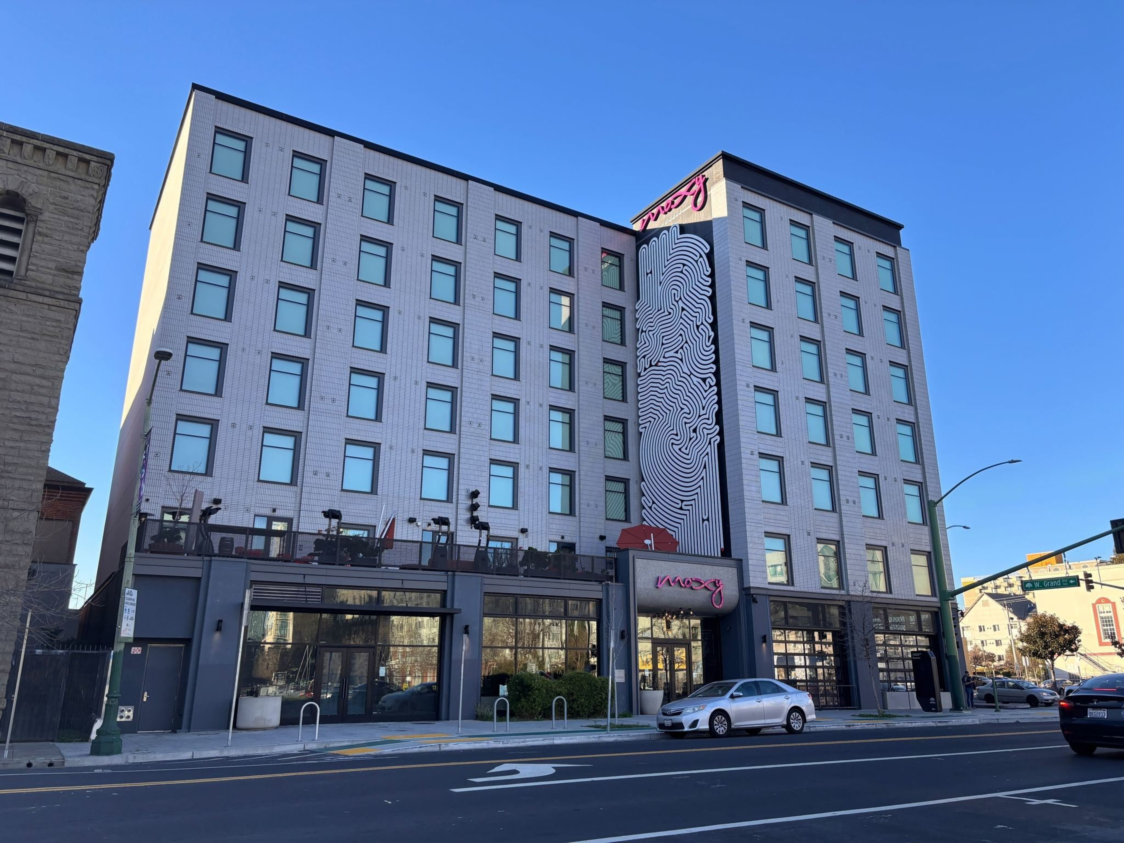 A six-story modern building with large windows features a black-and-white abstract mural and a sign reading "Moxy" above the entrance, with cars parked outside.