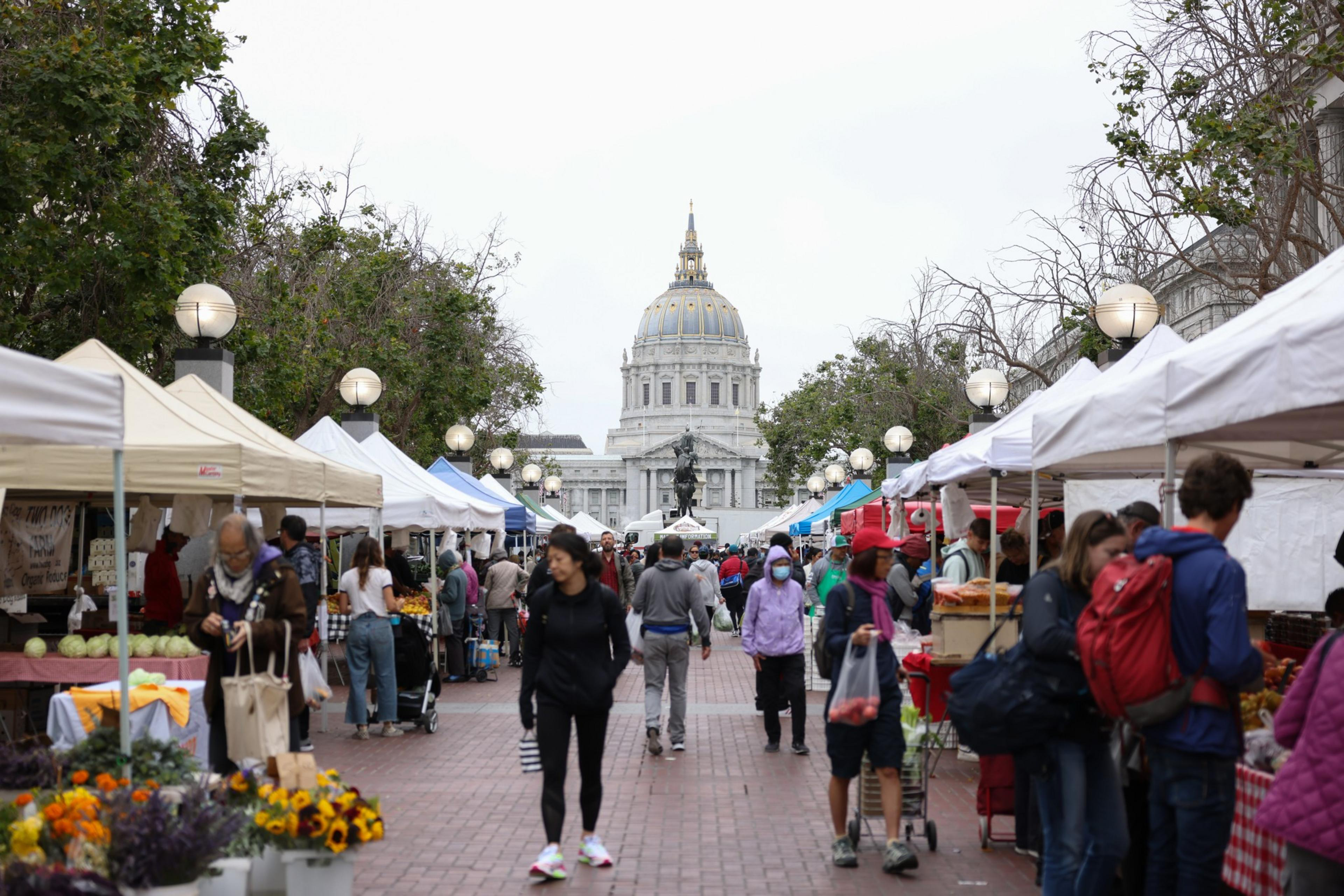 People browse a bustling outdoor market with white tents, located on a brick-paved pathway. A large domed building stands prominently in the background.