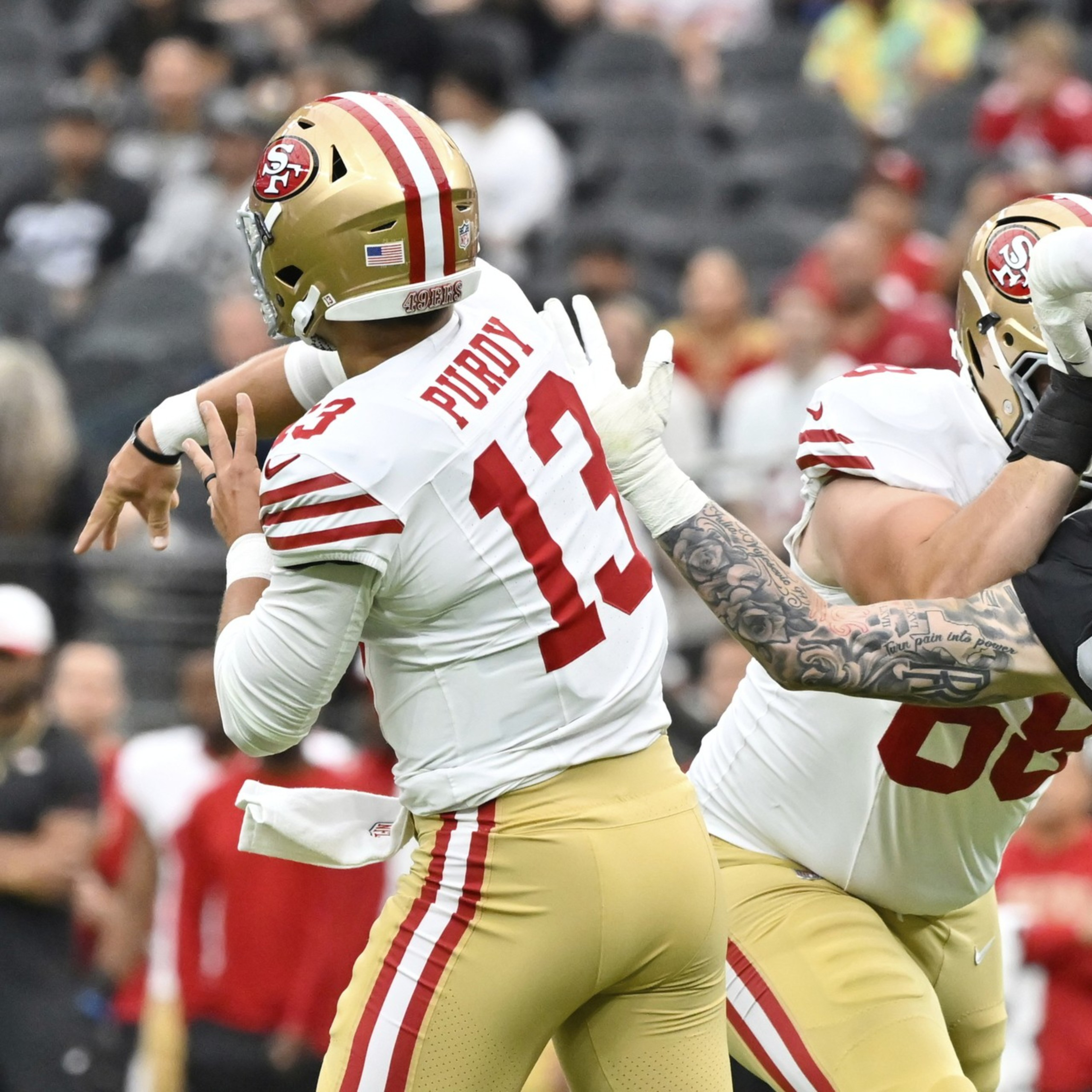A San Francisco 49ers quarterback wearing number 13 prepares to throw while a defender in black tries to block, and an offensive lineman blocks the defender.