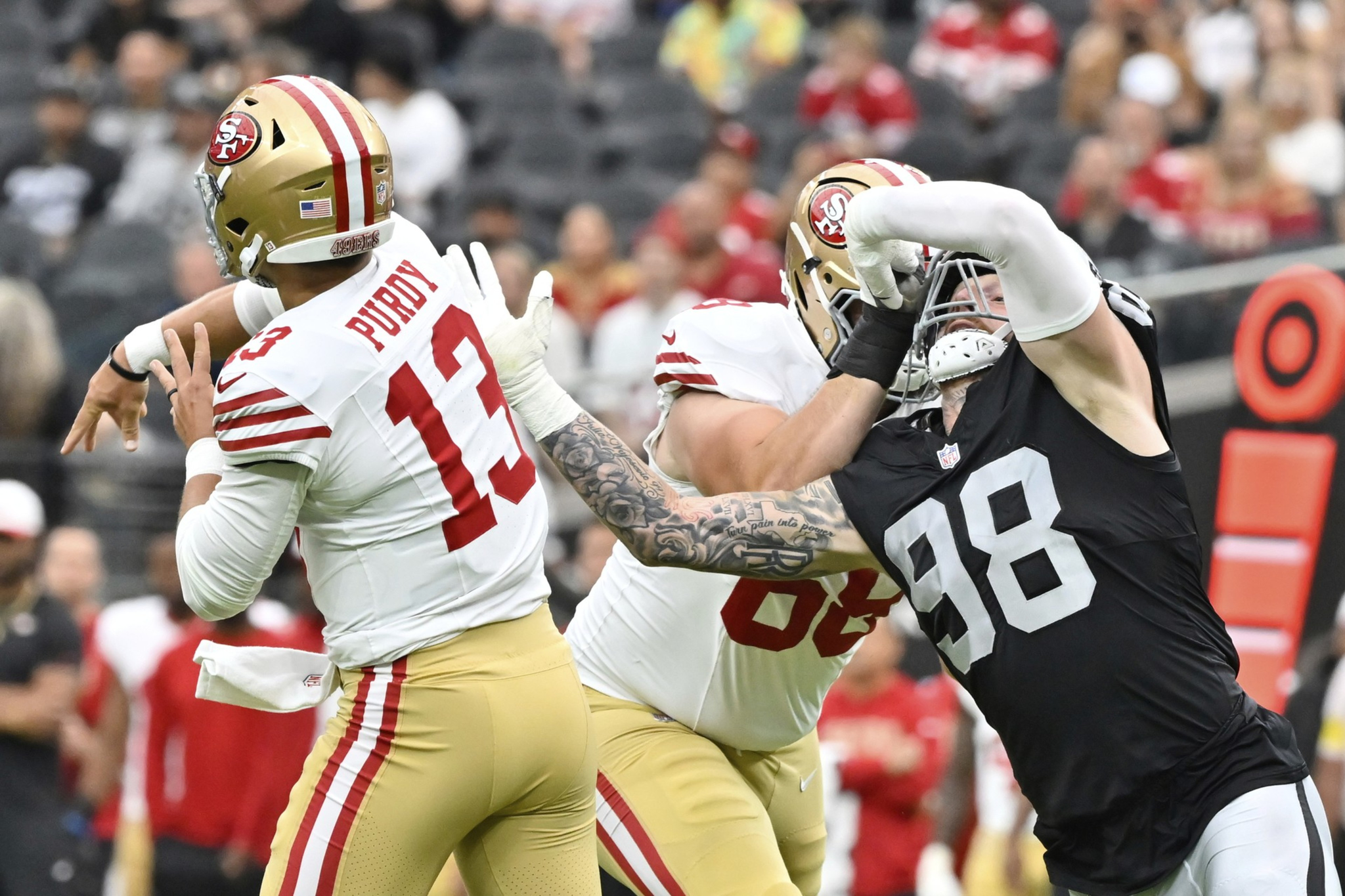 A San Francisco 49ers quarterback wearing number 13 prepares to throw while a defender in black tries to block, and an offensive lineman blocks the defender.