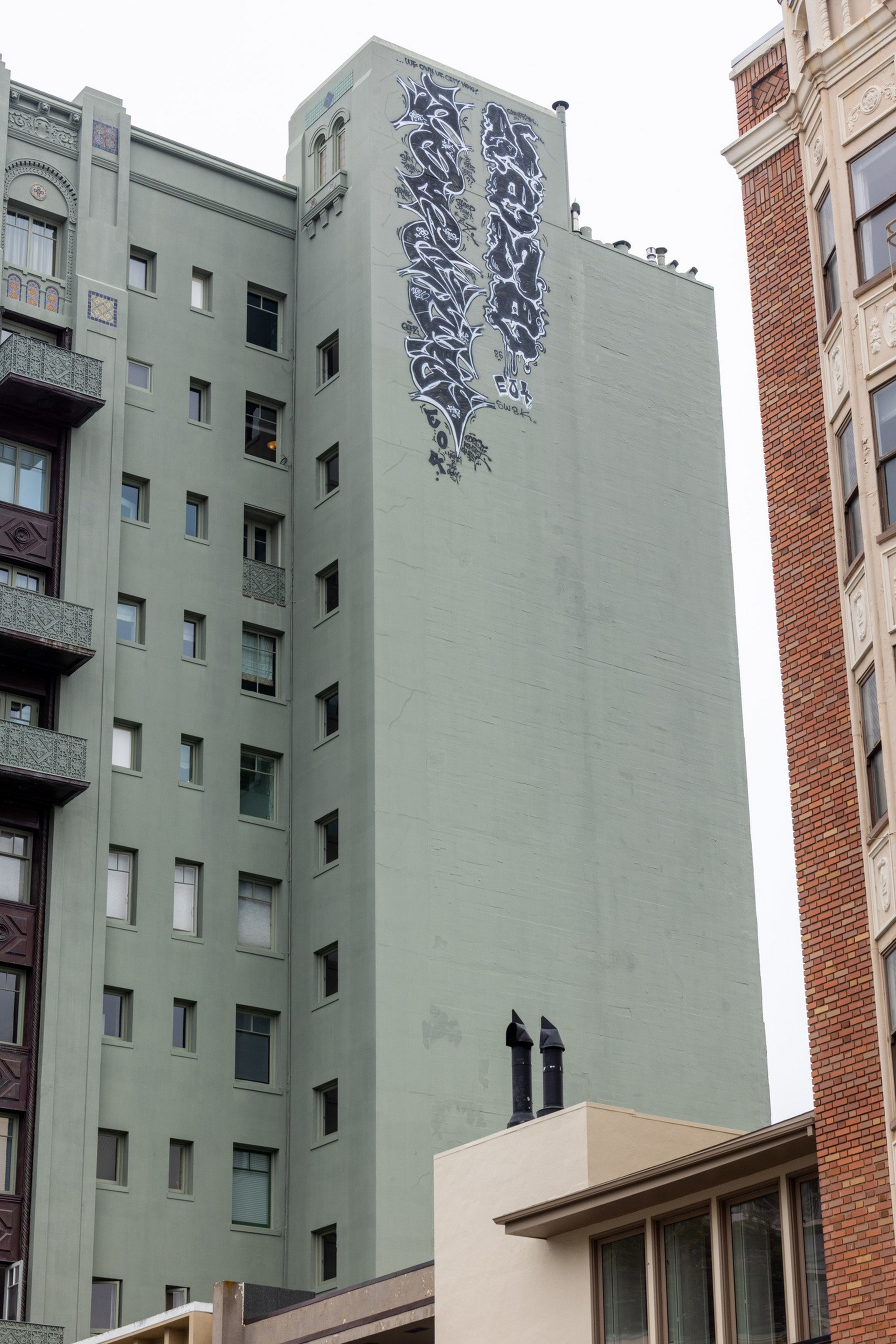 A tall greenish-gray building has black and white graffiti art of abstract shapes near its top, surrounded by other older-style buildings.
