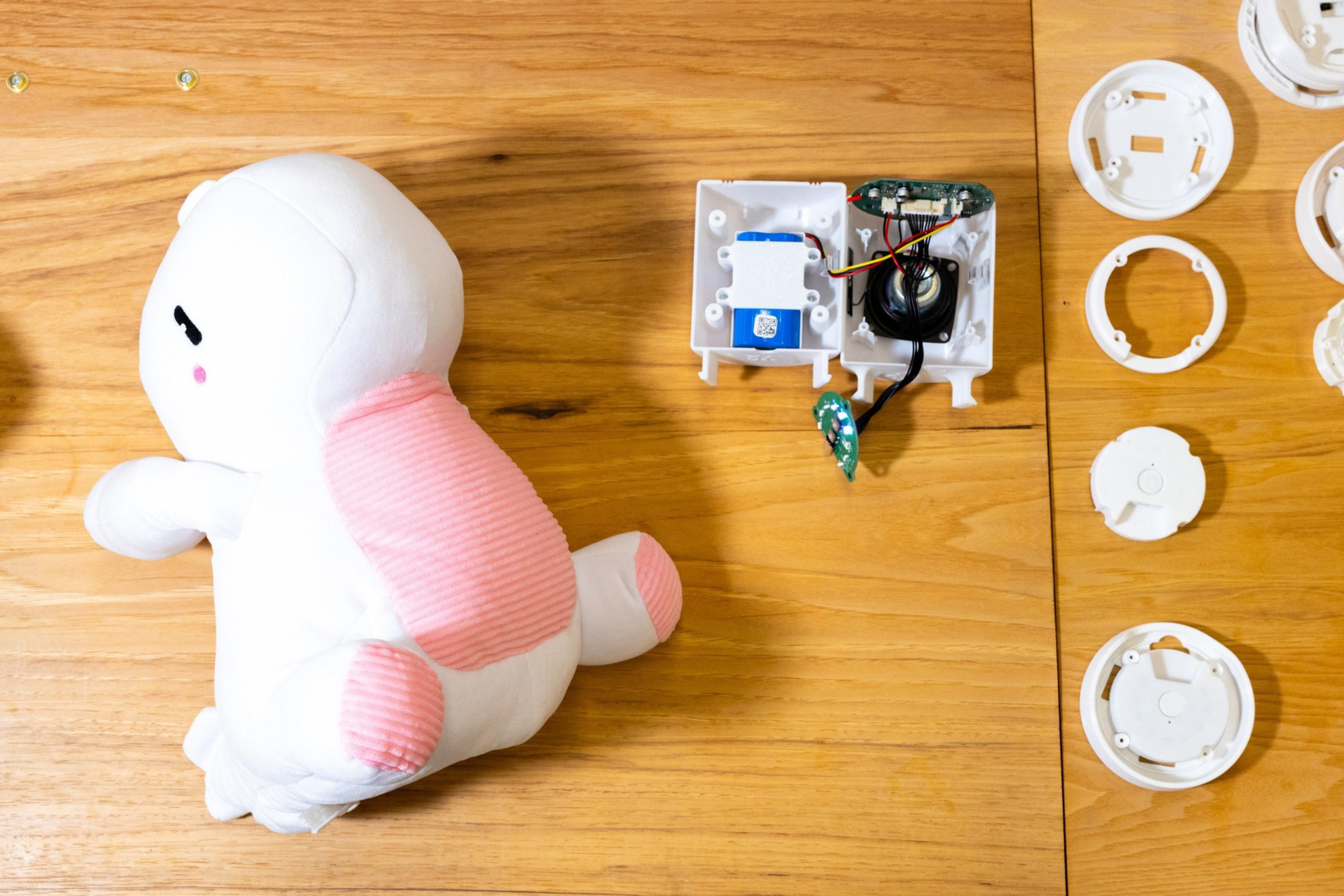 A white and pink stuffed animal is next to an open electronic device case with visible circuits and several white plastic circular parts on a wooden surface.