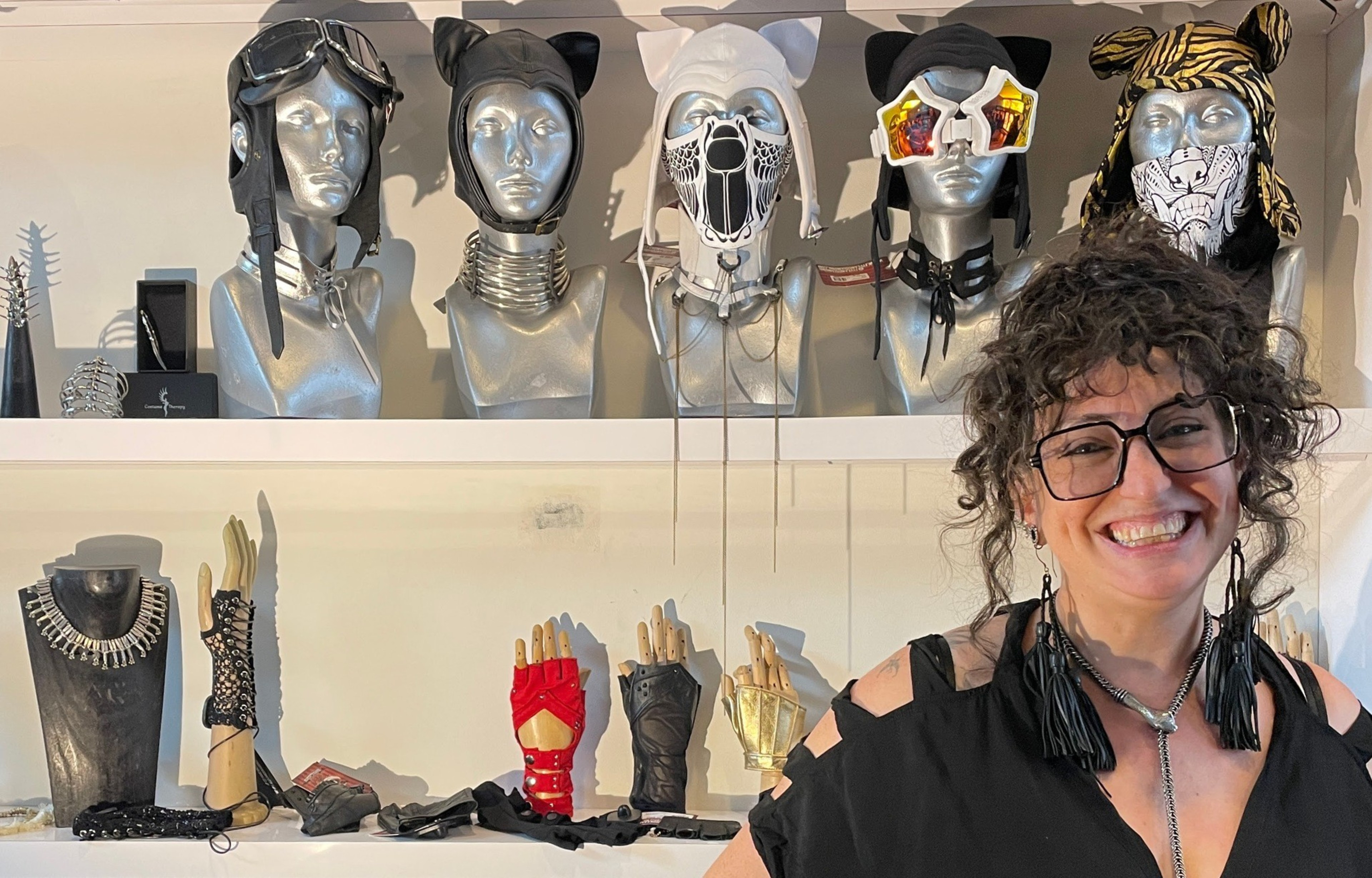 A woman with curly hair and glasses smiles in front of shelves displaying mannequins with unique headwear and accessories, including various masks, hats, and gloves.