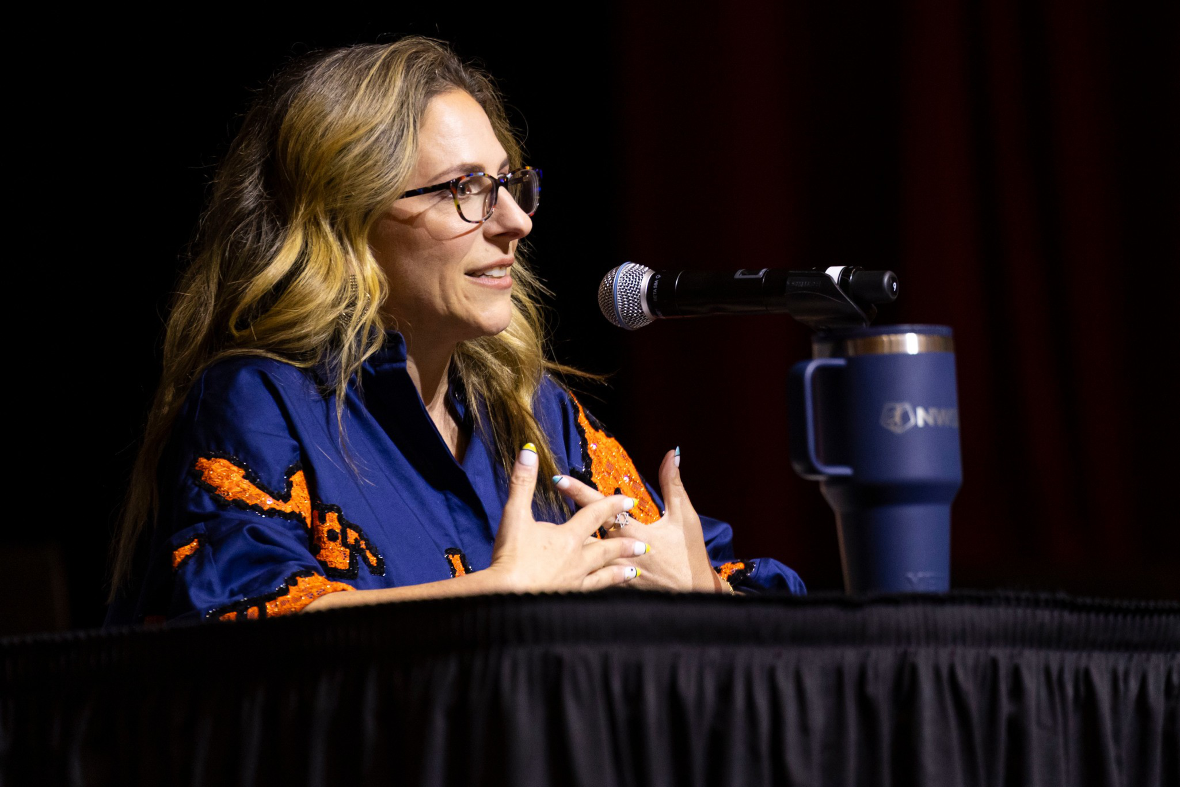 A woman with long blonde hair and glasses speaks into a microphone at a table, wearing a blue shirt with orange patterns, with a blue travel mug nearby.