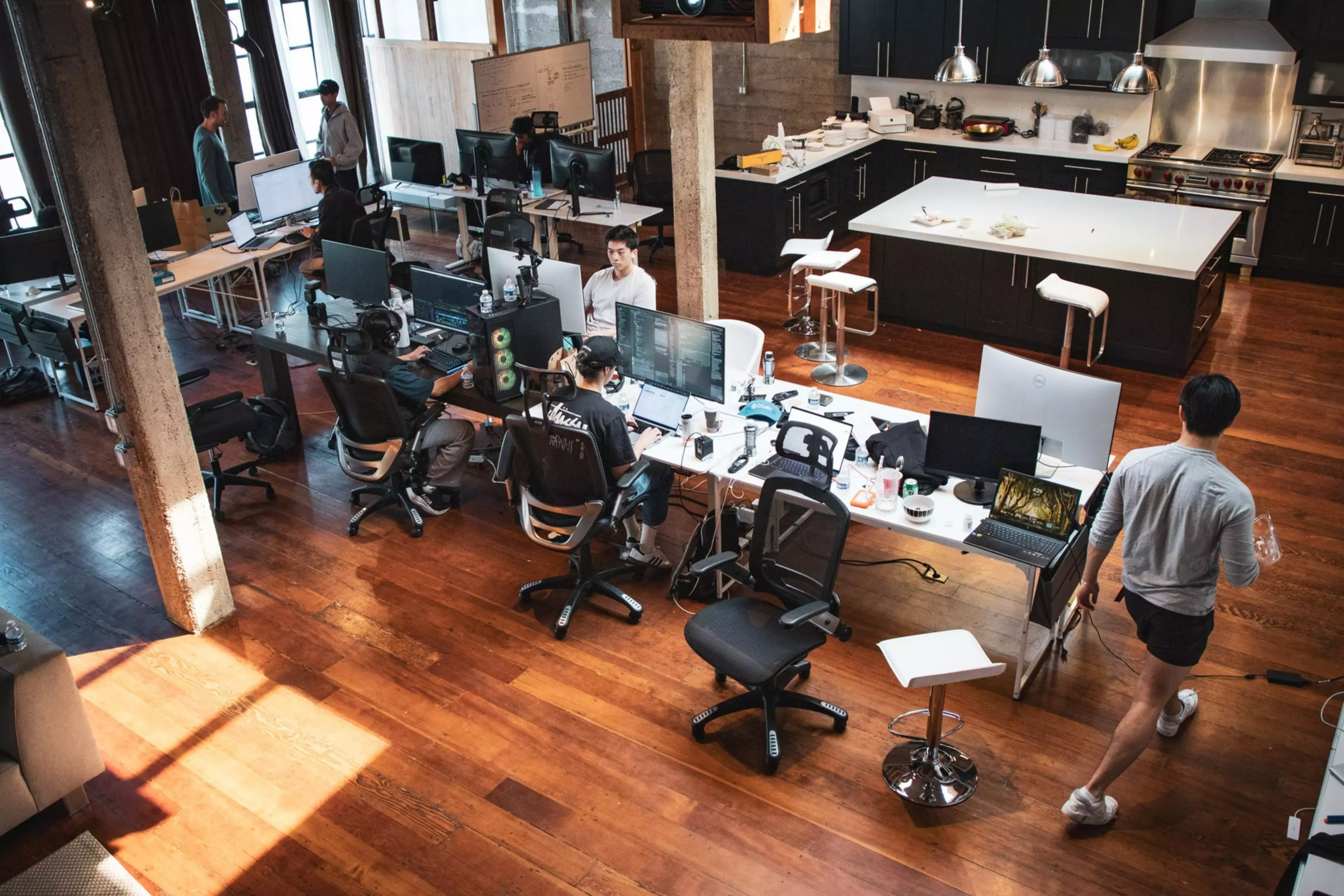 An open-plan office space features people working at desks with multiple computers, a kitchen area in the back, and a wooden floor throughout.