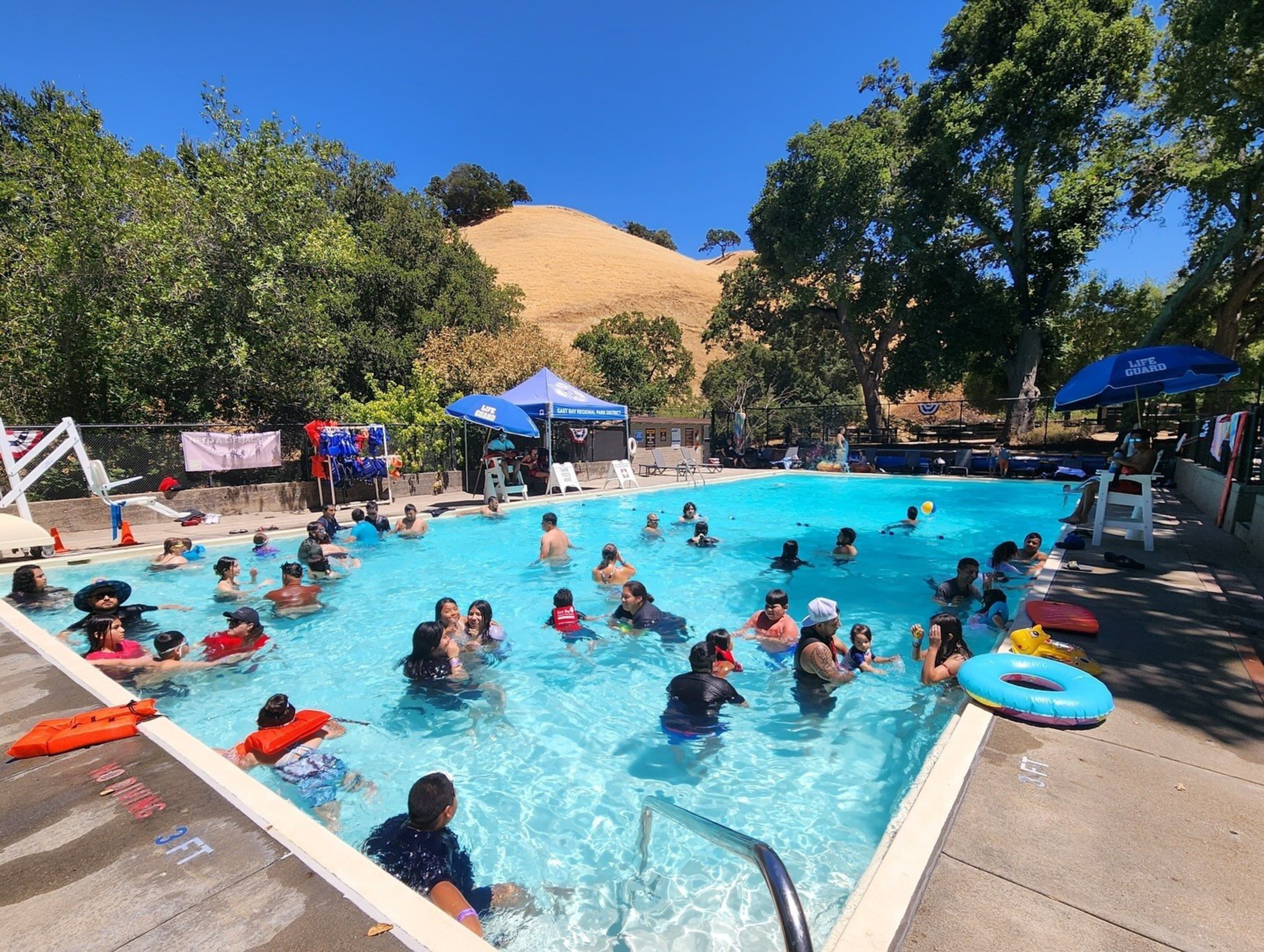 A crowded outdoor swimming pool with people enjoying the water under sunny skies. Lifeguard chairs and colorful umbrellas are around the poolside.