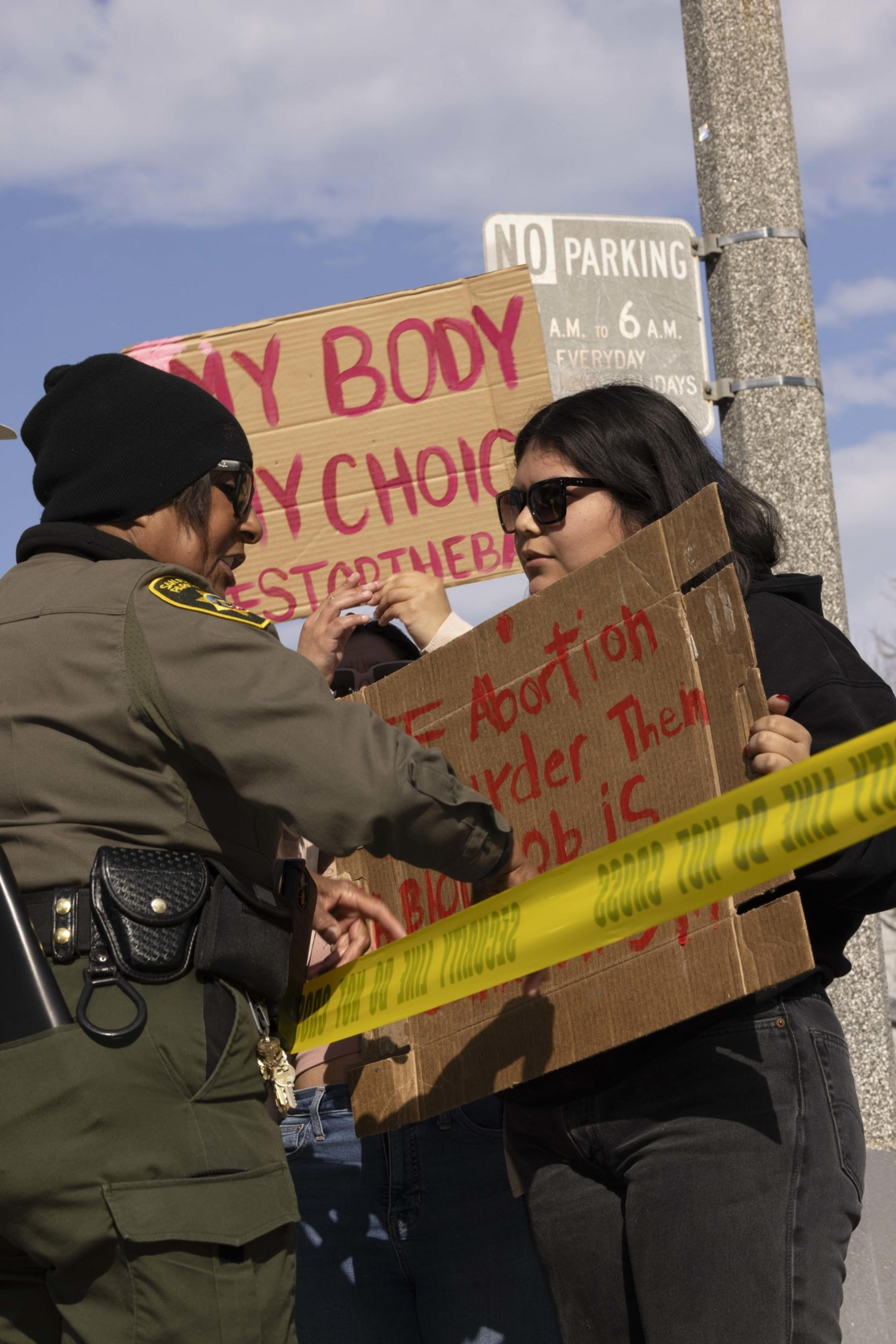 A police officer and a protester holding signs engage behind yellow tape. The signs display messages about body autonomy and abortion rights.