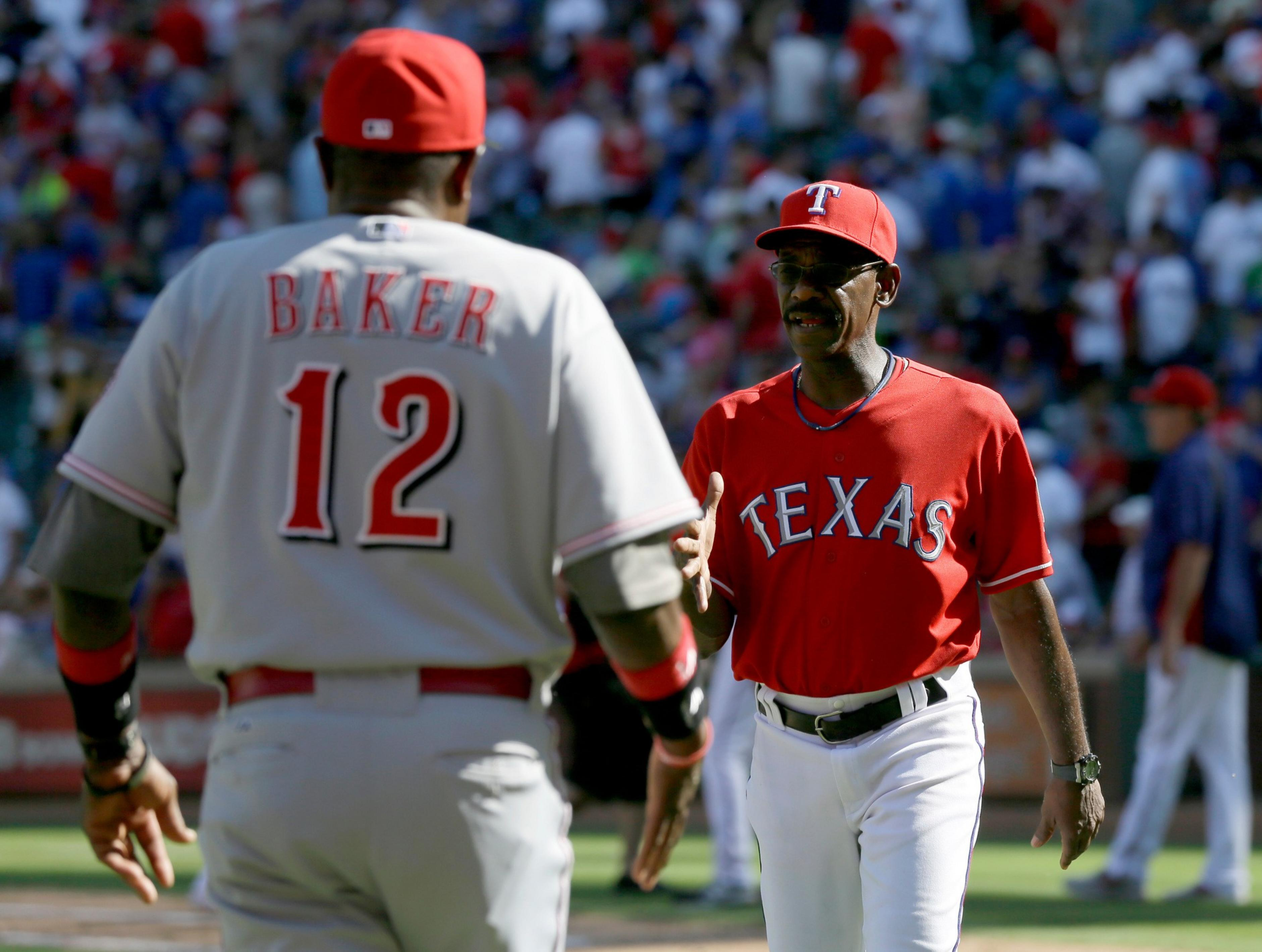 A baseball player in a gray uniform with “Baker” and number 12 faces a man in a red Texas Rangers shirt and cap, extending a handshake.