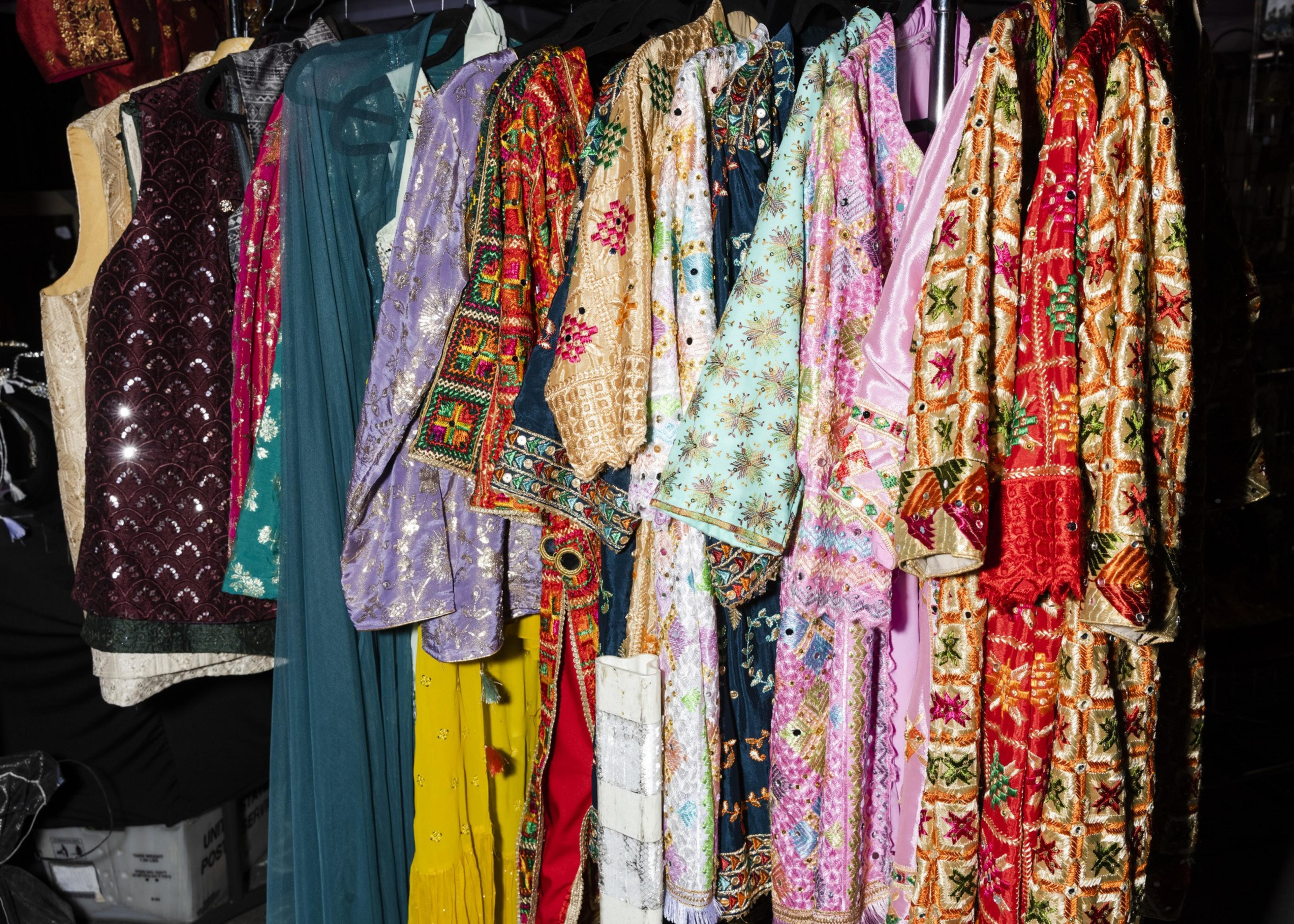 The image shows a row of colorful, intricately embroidered clothes hanging on a rack. The fabrics are vibrant, featuring reds, greens, blues, and gold designs.