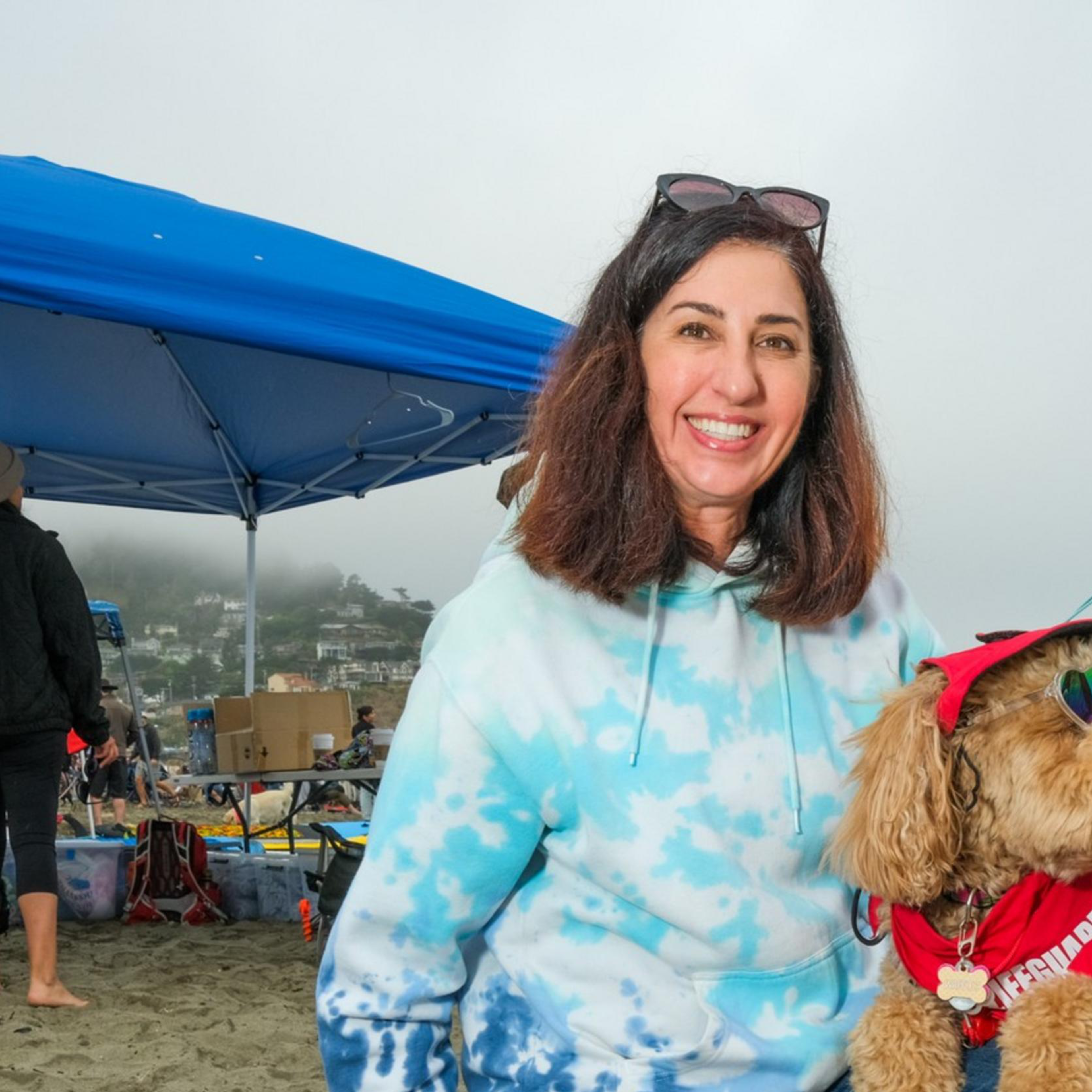 A smiling woman in a blue tie-dye hoodie kneels on a beach next to a dog wearing sunglasses and a red bandana. Beachgoers and tents are in the background.