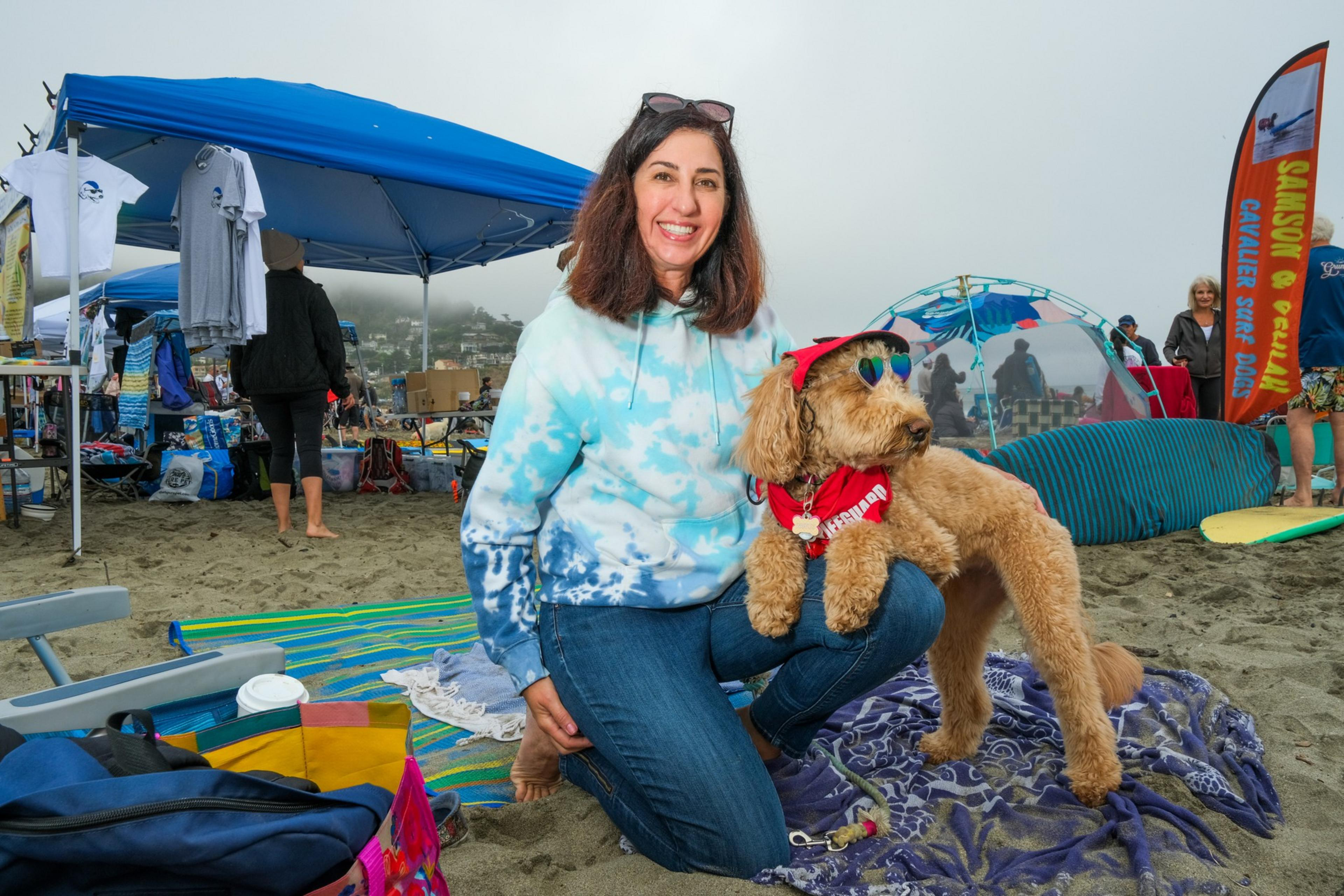 A smiling woman in a blue tie-dye hoodie kneels on a beach next to a dog wearing sunglasses and a red bandana. Beachgoers and tents are in the background.