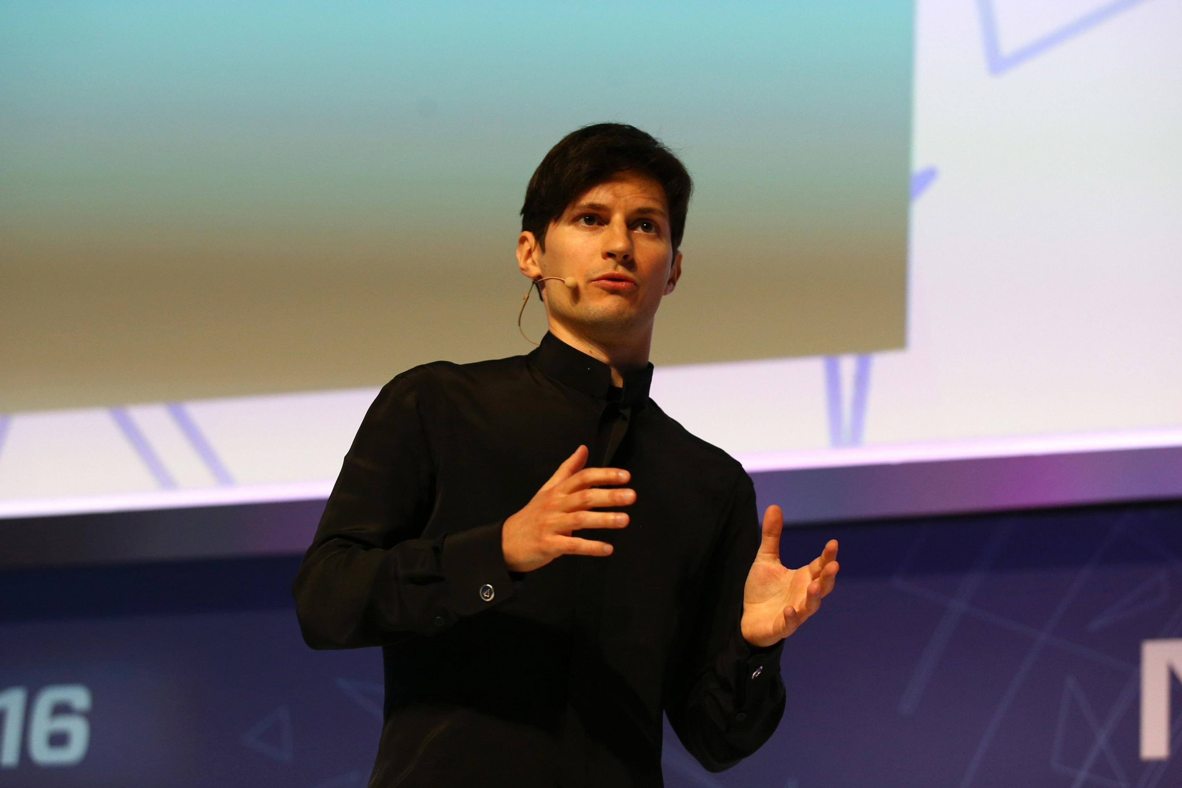 A man in a black shirt speaks at a podium, gesturing mid-speech with a projected blue background.