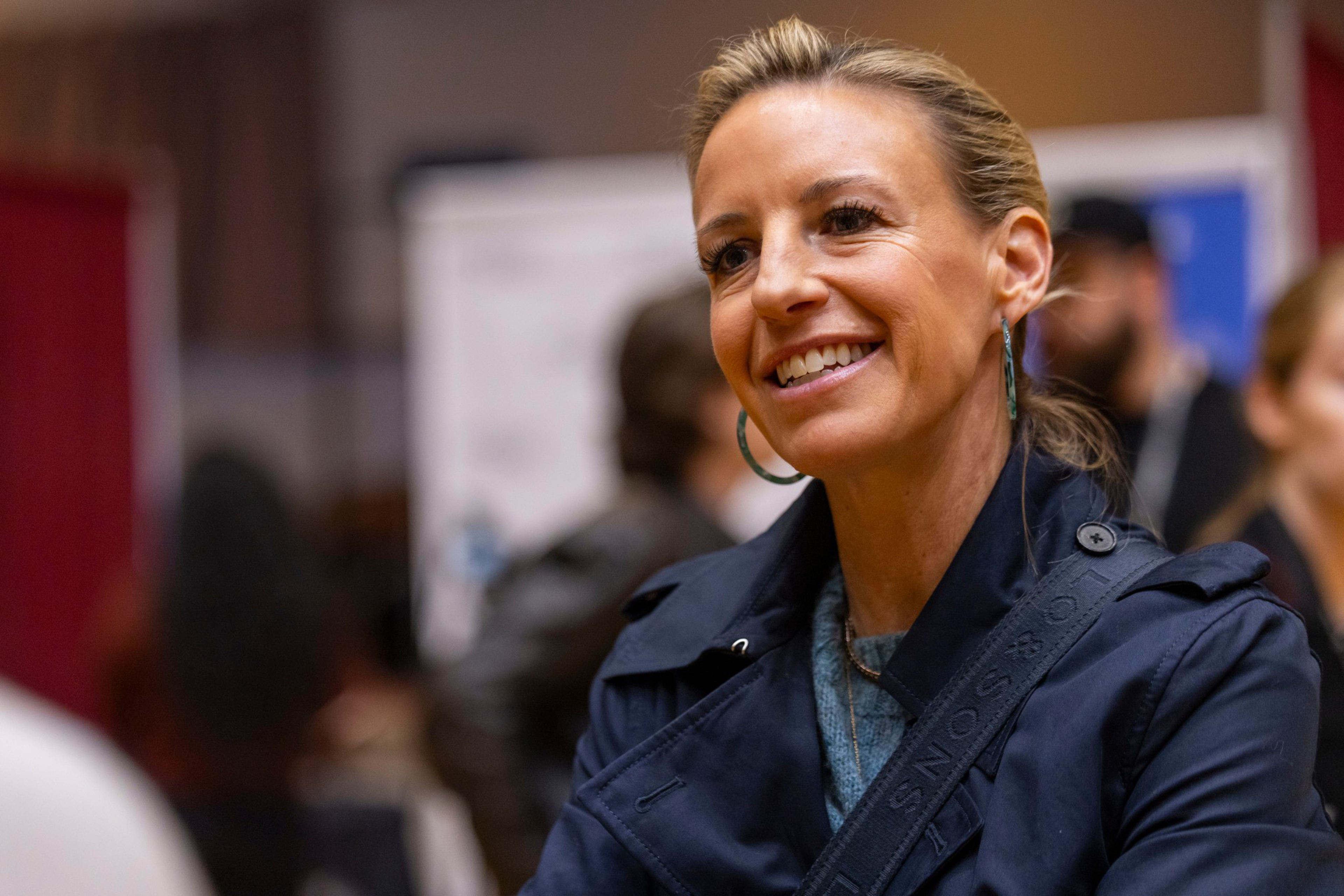 A woman with blonde hair pulled back is smiling and wearing a dark jacket and hoop earrings in a softly lit indoor setting with blurred people in the background.