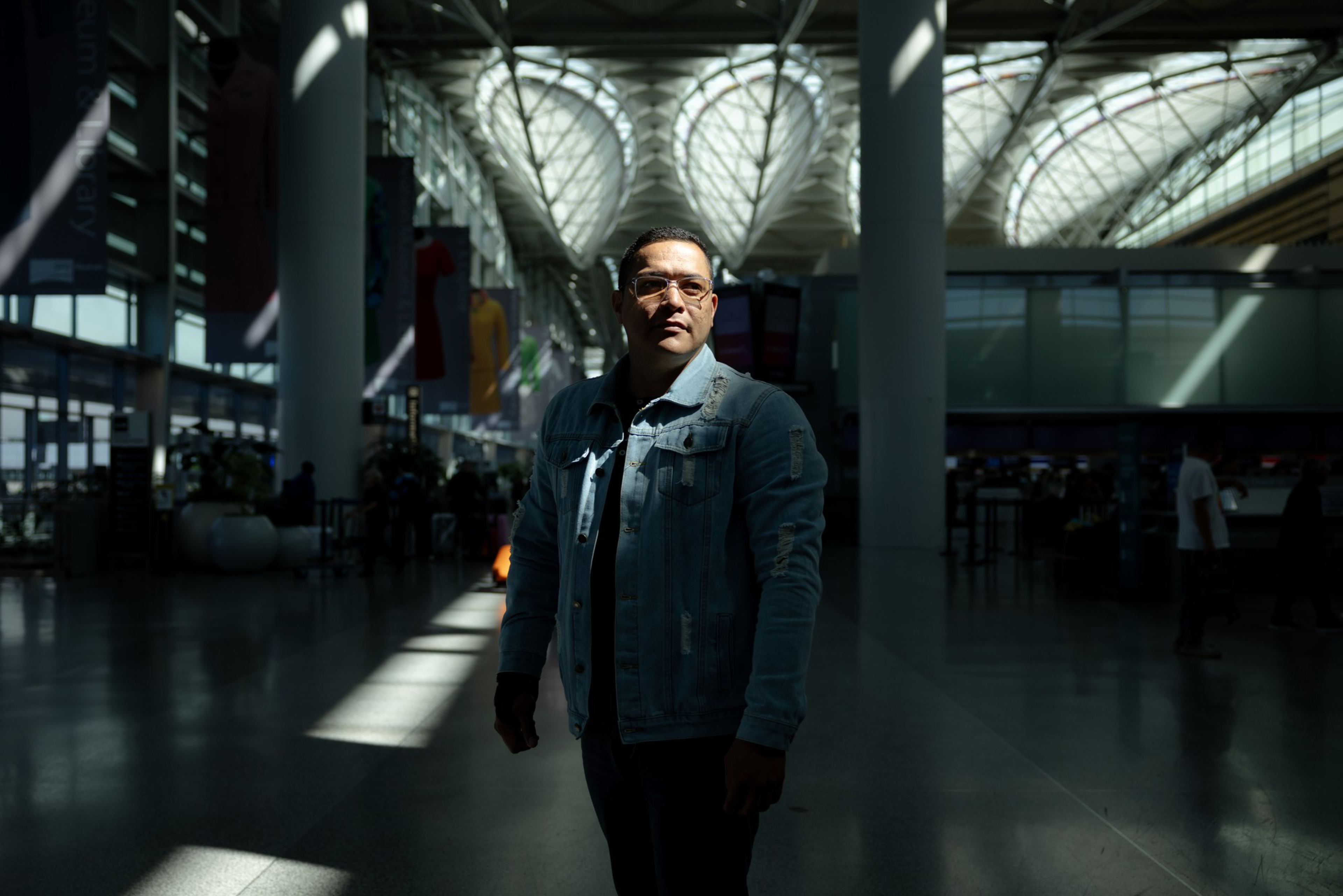 A man wearing glasses and a denim jacket stands in a large, sunlit indoor space with high ceilings and scattered shadows.