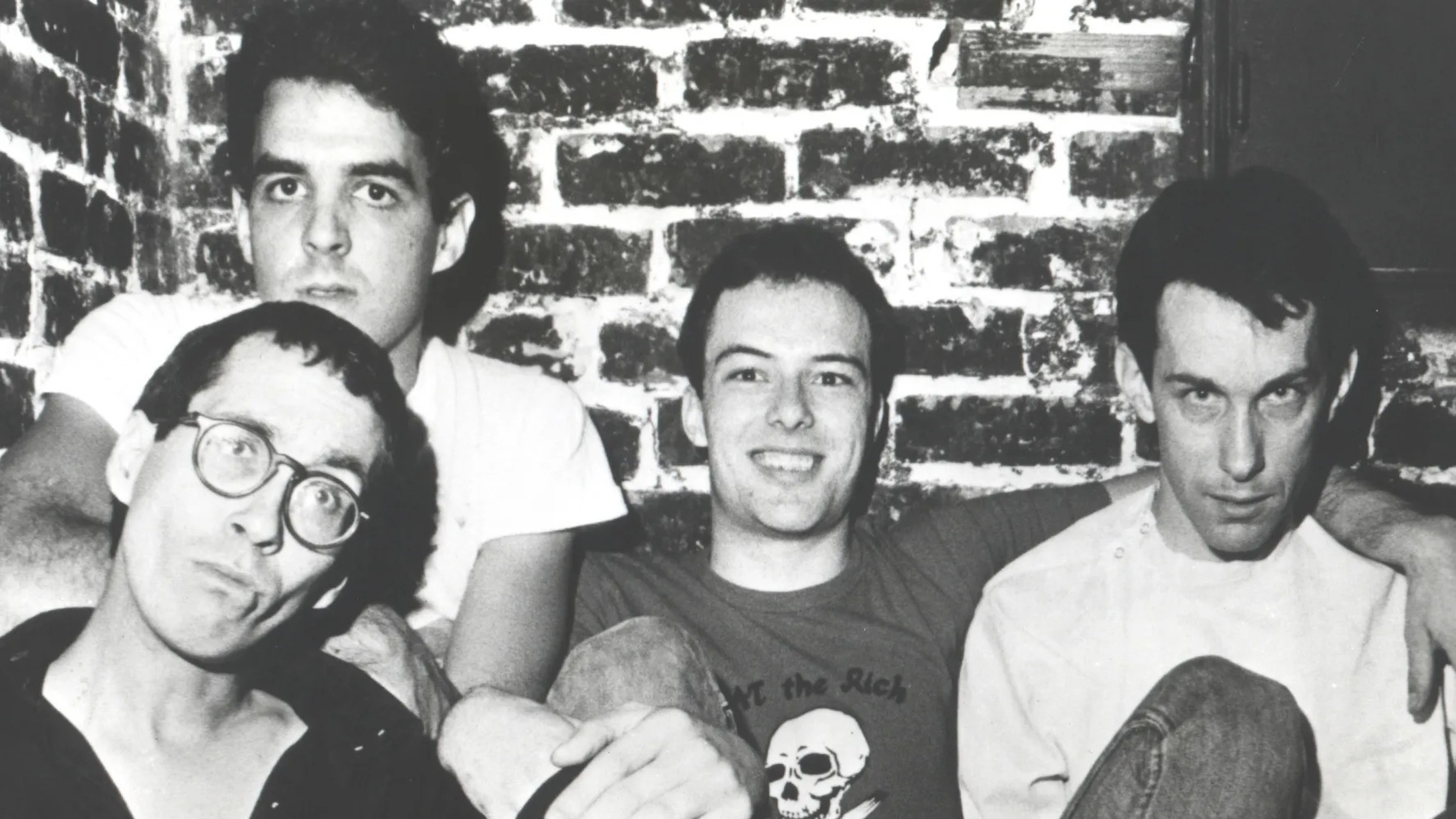 Four men sit closely together against a rough brick wall, three looking serious or neutral and one smiling while wearing a skull-printed shirt.