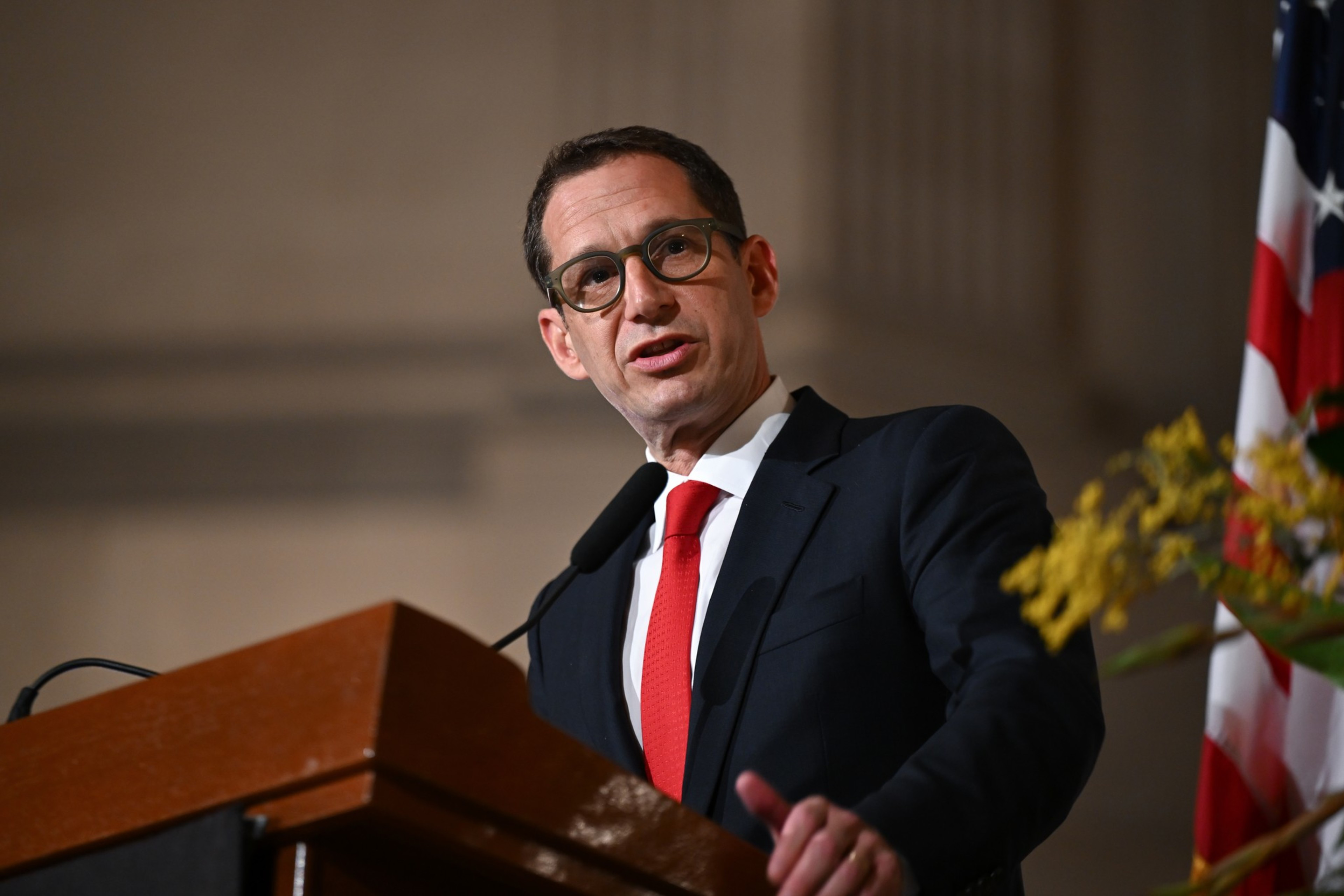 A man in a dark suit, white shirt, and red tie is speaking at a wooden podium with a microphone, with an American flag and flowers nearby.