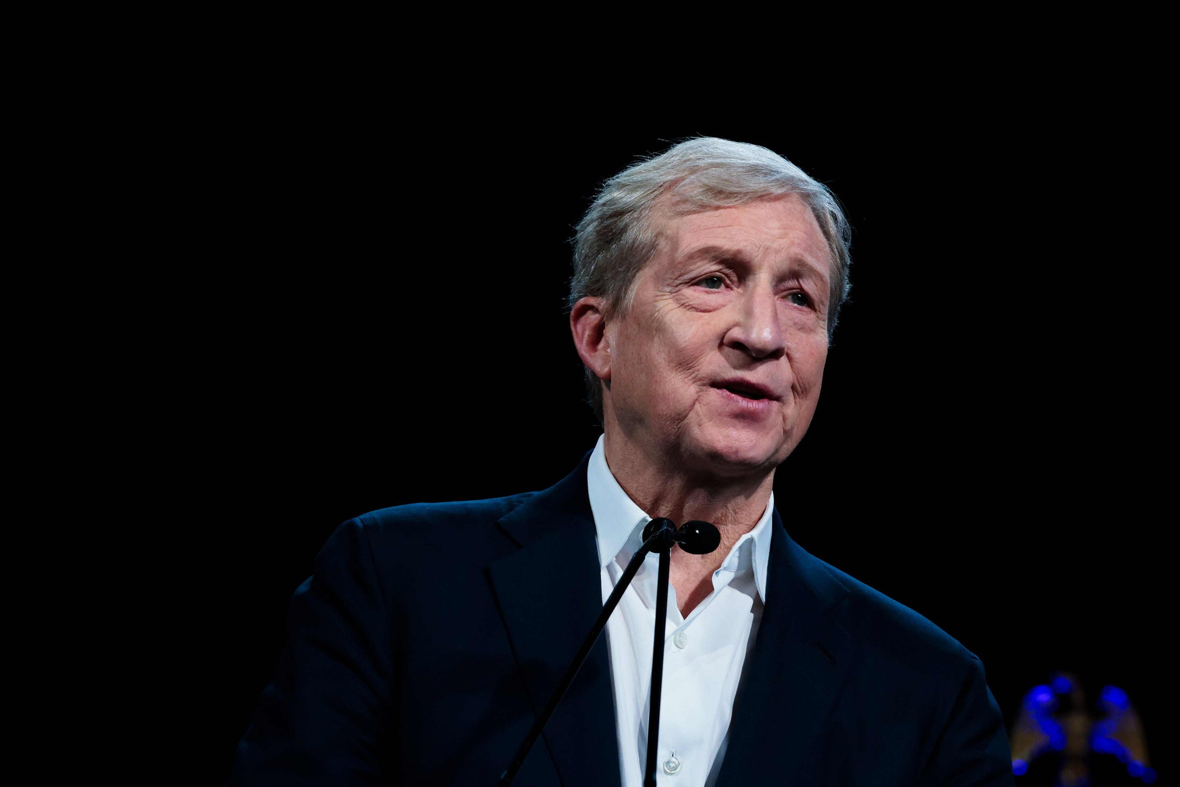 An older man with gray hair wears a dark suit and white shirt, speaking into a microphone against a black background.
