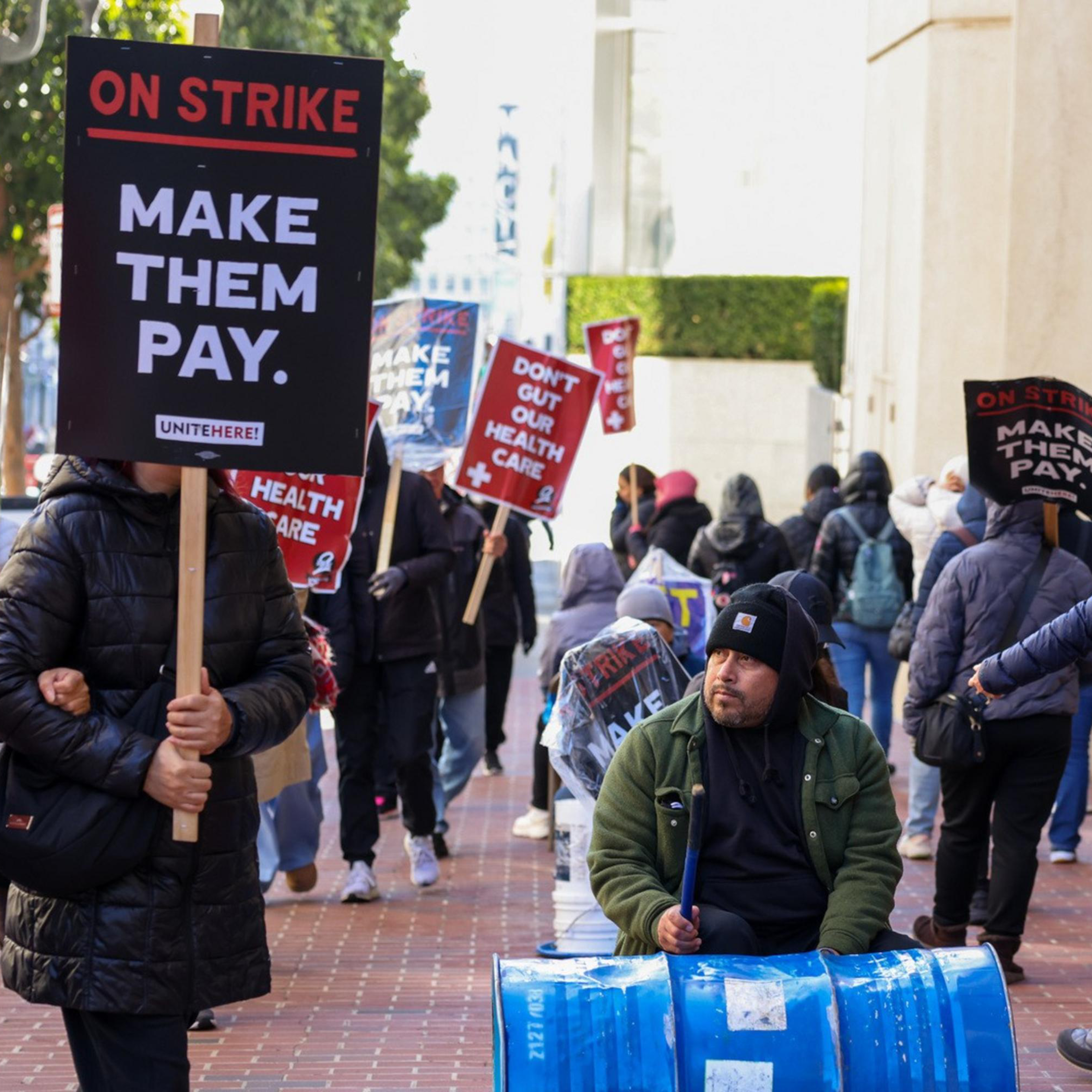 Protesters stand on a brick sidewalk holding signs that say "DON'T GUT OUR HEALTH CARE" and "MAKE THEM PAY," with a man drumming on a blue barrel.