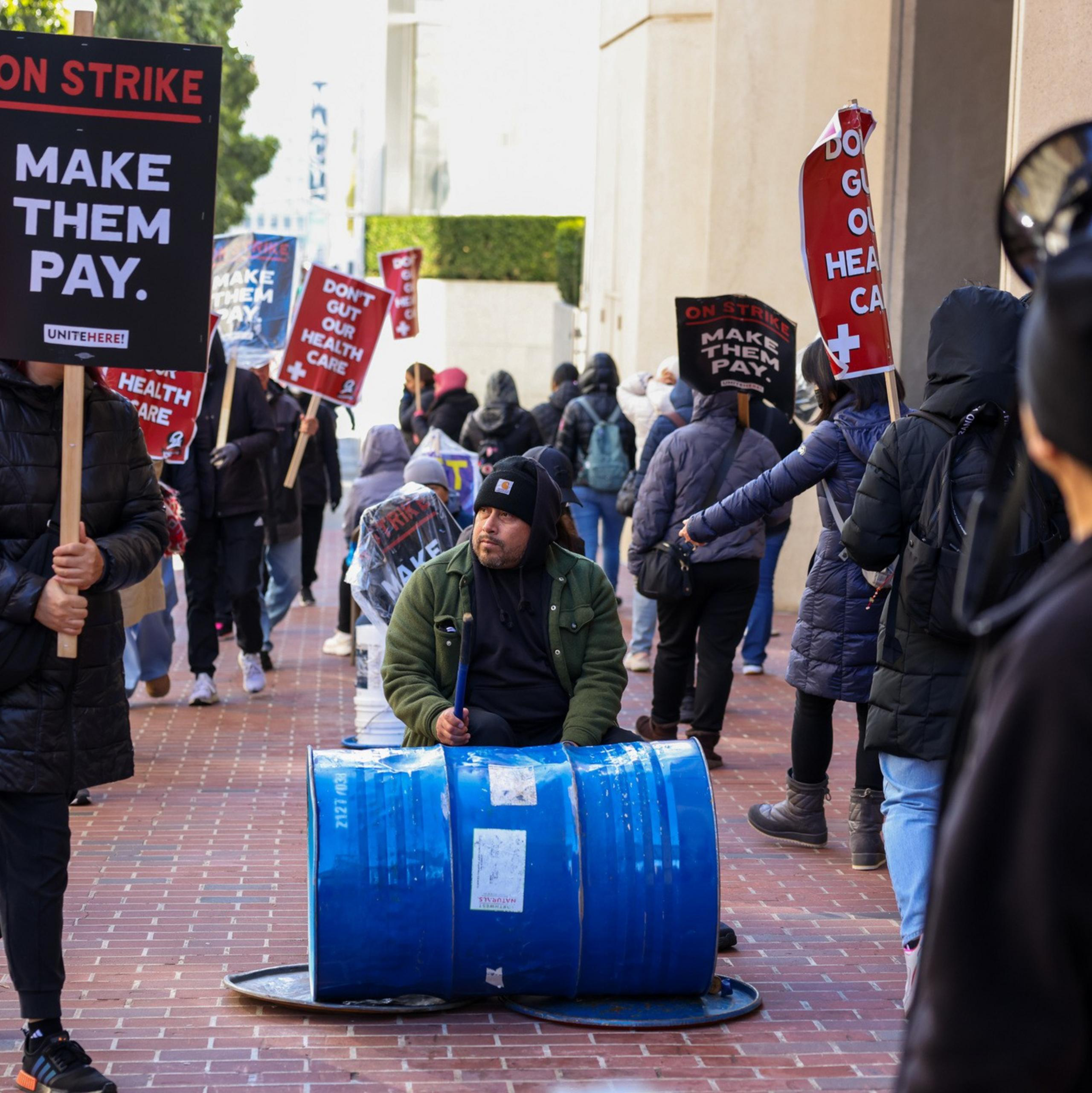 Protesters stand on a brick sidewalk holding signs that say "DON'T GUT OUR HEALTH CARE" and "MAKE THEM PAY," with a man drumming on a blue barrel.