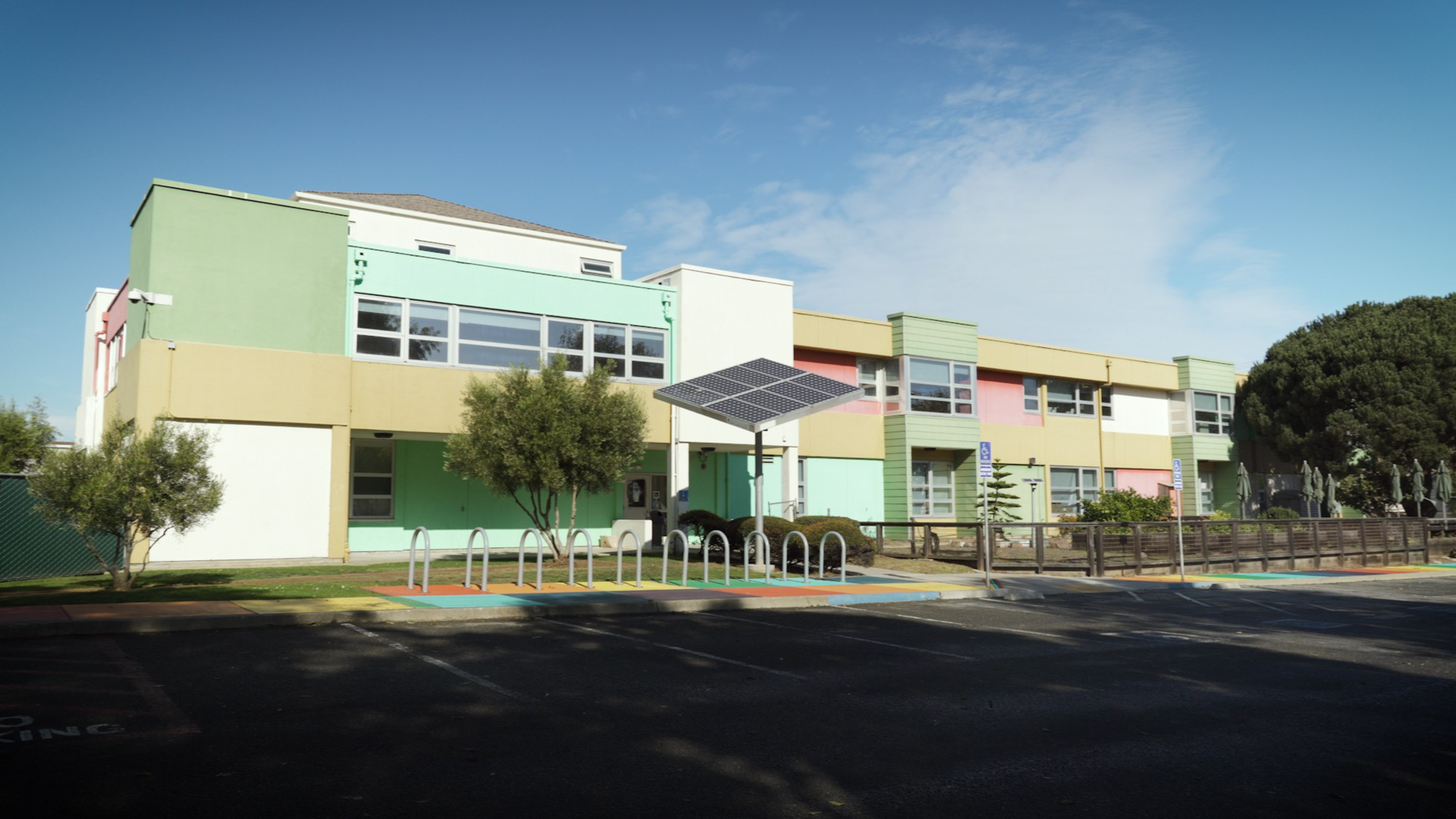A two-story building with green, yellow, and pink walls stands under a blue sky, featuring trees, bike racks, and a solar panel canopy in front.
