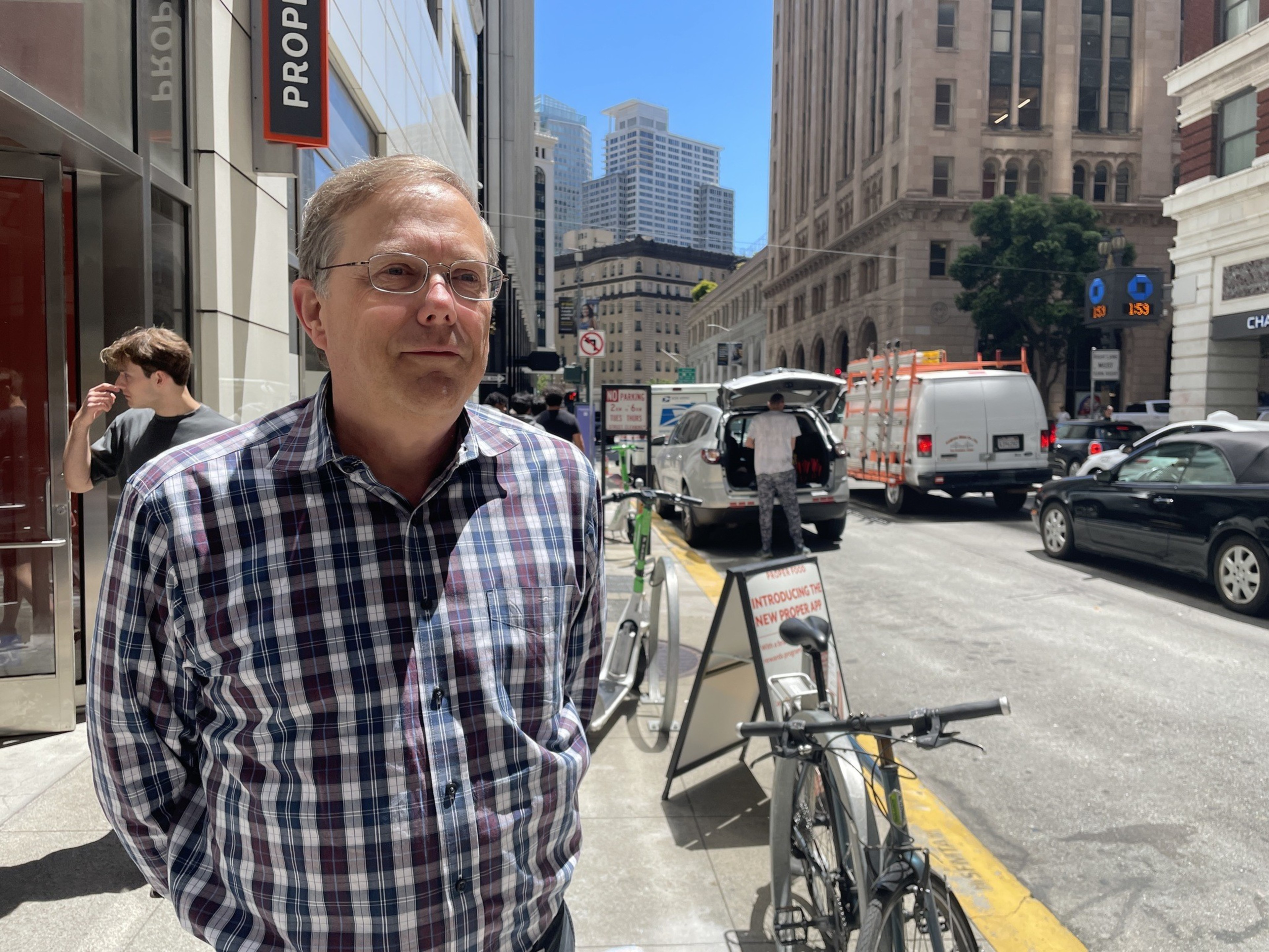 A man in a plaid shirt stands on a bustling city street with several vehicles and bicycles nearby. Tall buildings and a bright blue sky are in the background.