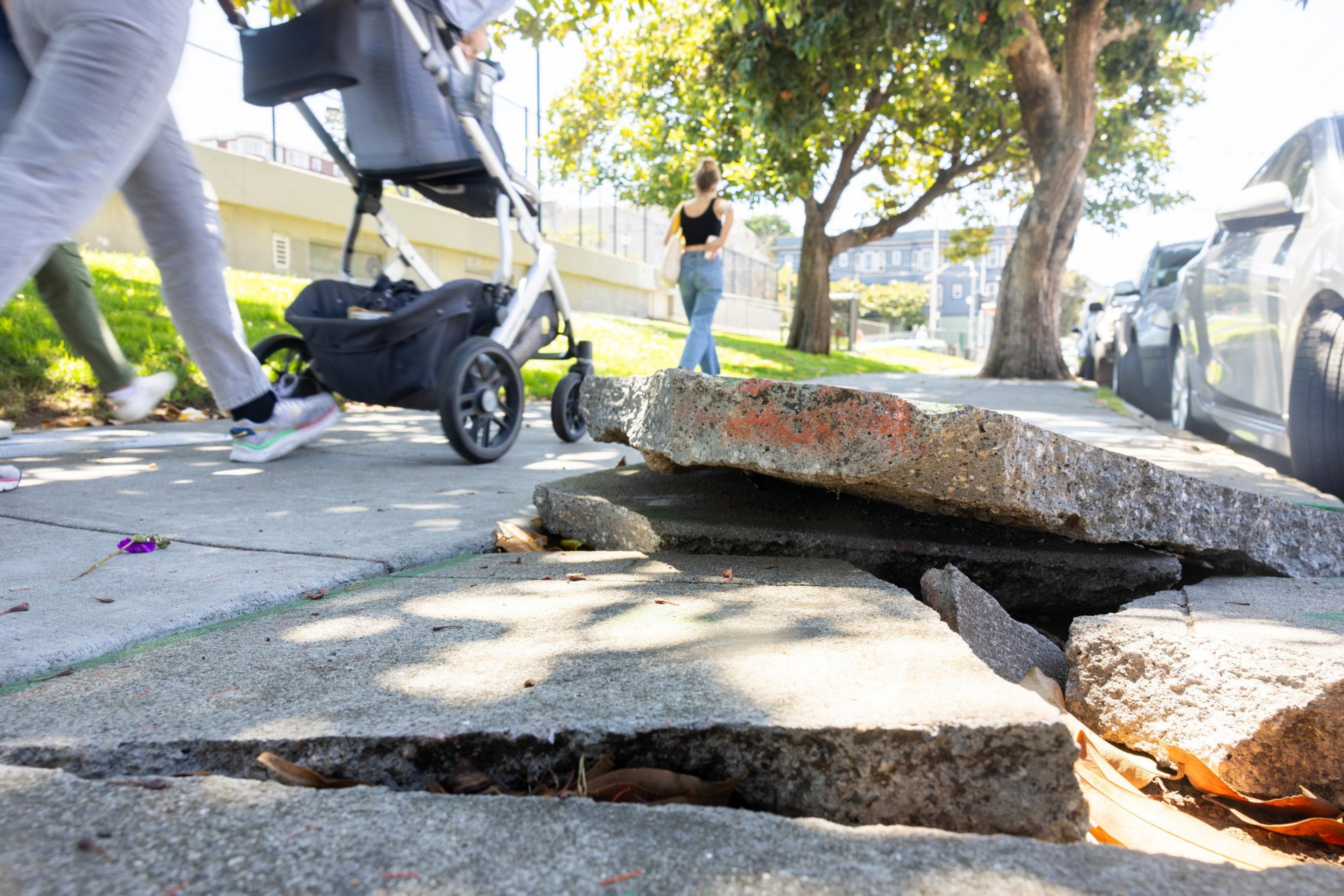 A sidewalk with broken, uneven concrete slabs. Nearby, a person pushes a stroller while another person walks in the background. Trees and cars are visible.
