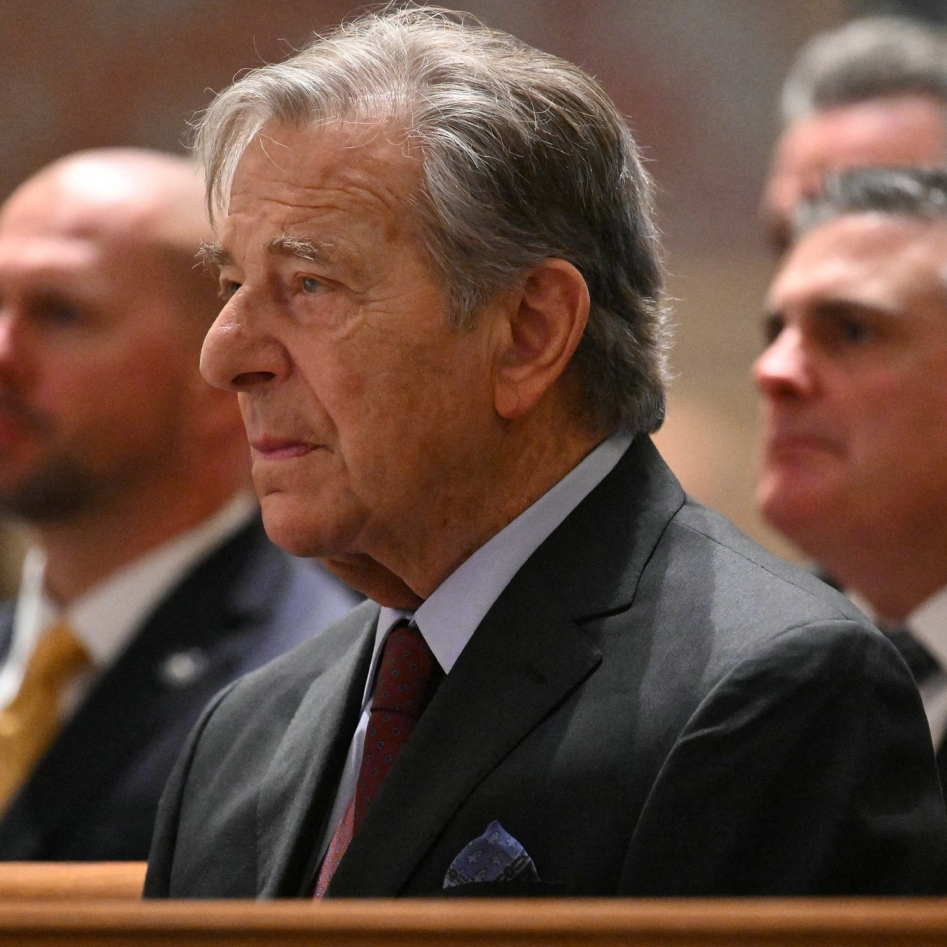 A man with gray hair is seated, wearing a dark suit and tie, looking ahead thoughtfully. Other similarly dressed individuals are visible but blurred in the background.