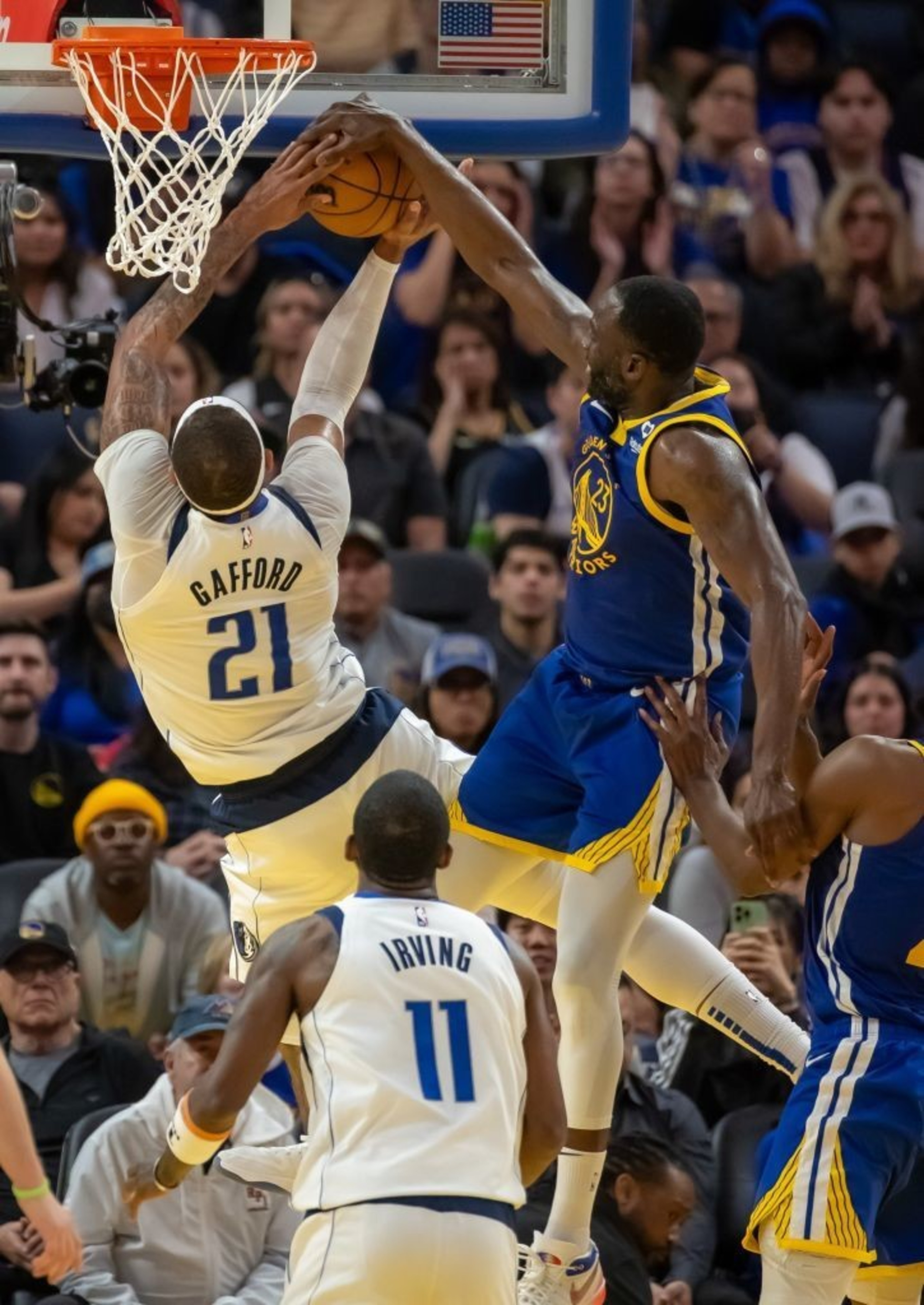 Two basketball players are mid-air near the hoop. The player in white attempts a dunk while a player in blue defends, reaching up to block the shot.